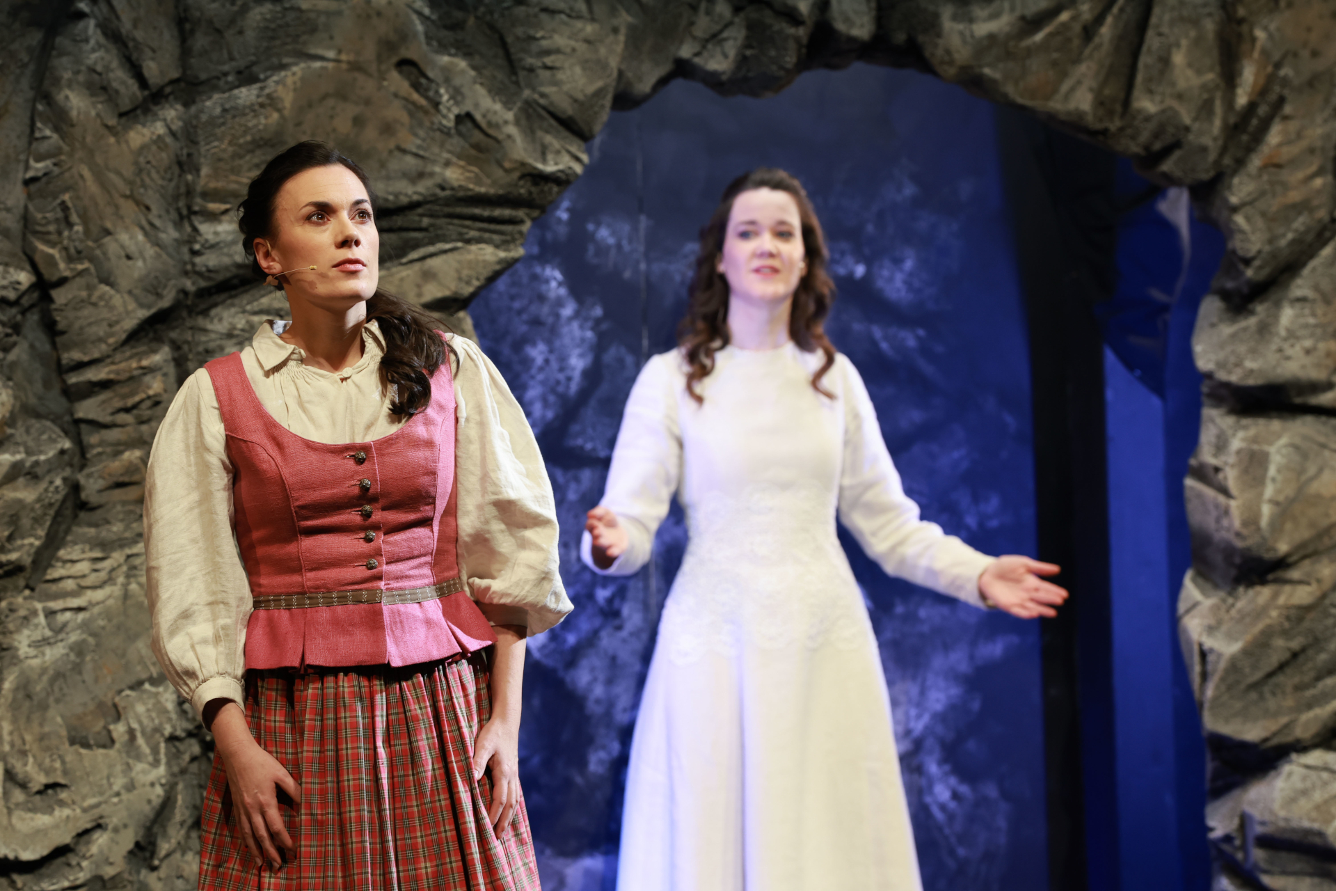 Zwei Frauen in historischen Kost&uuml;men treten auf der B&uuml;hne auf, eine in rosa Karo, die andere in wei&szlig;, eingerahmt von einem Felsenbogen.