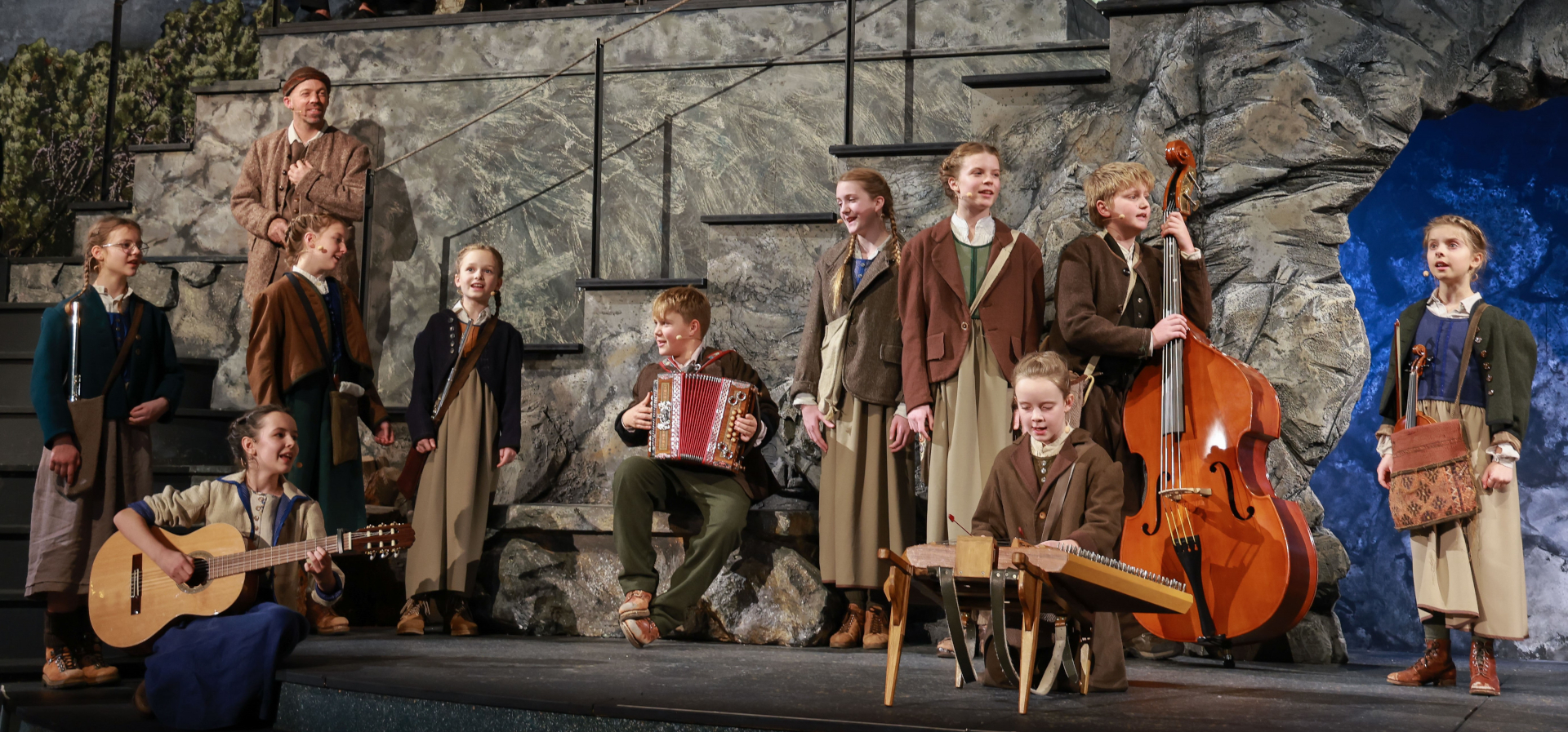Kinder in Kost&uuml;men spielen auf der B&uuml;hne mit Instrumenten, darunter Gitarre, Akkordeon und Kontrabass.