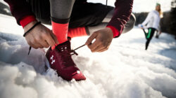 Eine Person bindet kastanienbraune Laufschuhe im Schnee, während sich eine andere Person im Hintergrund dehnt.