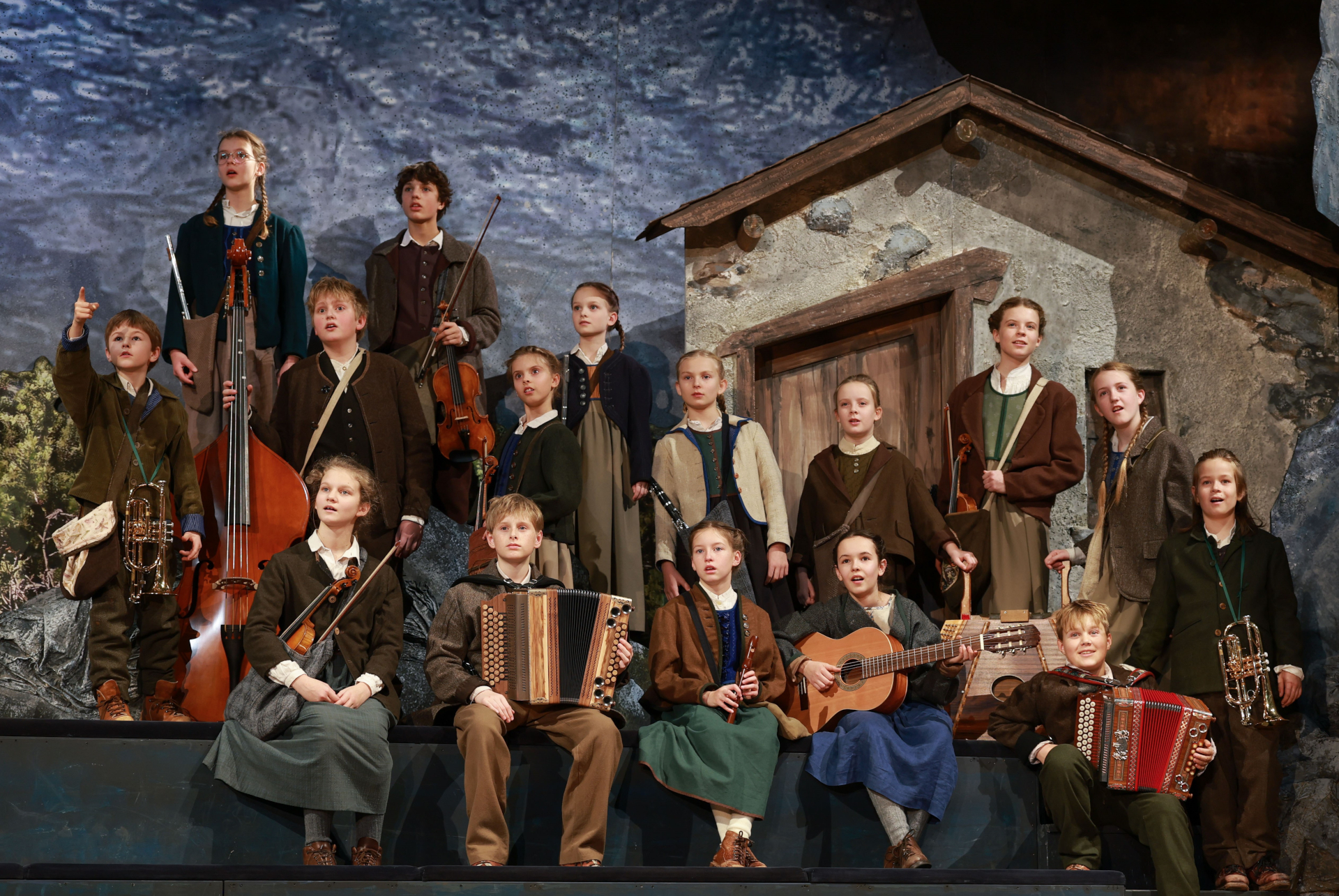 Eine Gruppe von Kindern in traditioneller &ouml;sterreichischer Kleidung spielt auf der B&uuml;hne vor einer rustikalen Hauskulisse Musikinstrumente.