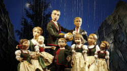 Puppen in traditioneller österreichischer Tracht spielen auf der Bühne mit Bergen und Bäumen im Hintergrund.