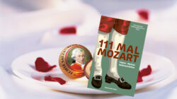 Eine Mozart-Schokolade und das Buch 111 Mal Mozart auf einem weißen Teller mit verstreuten roten Rosenblättern.