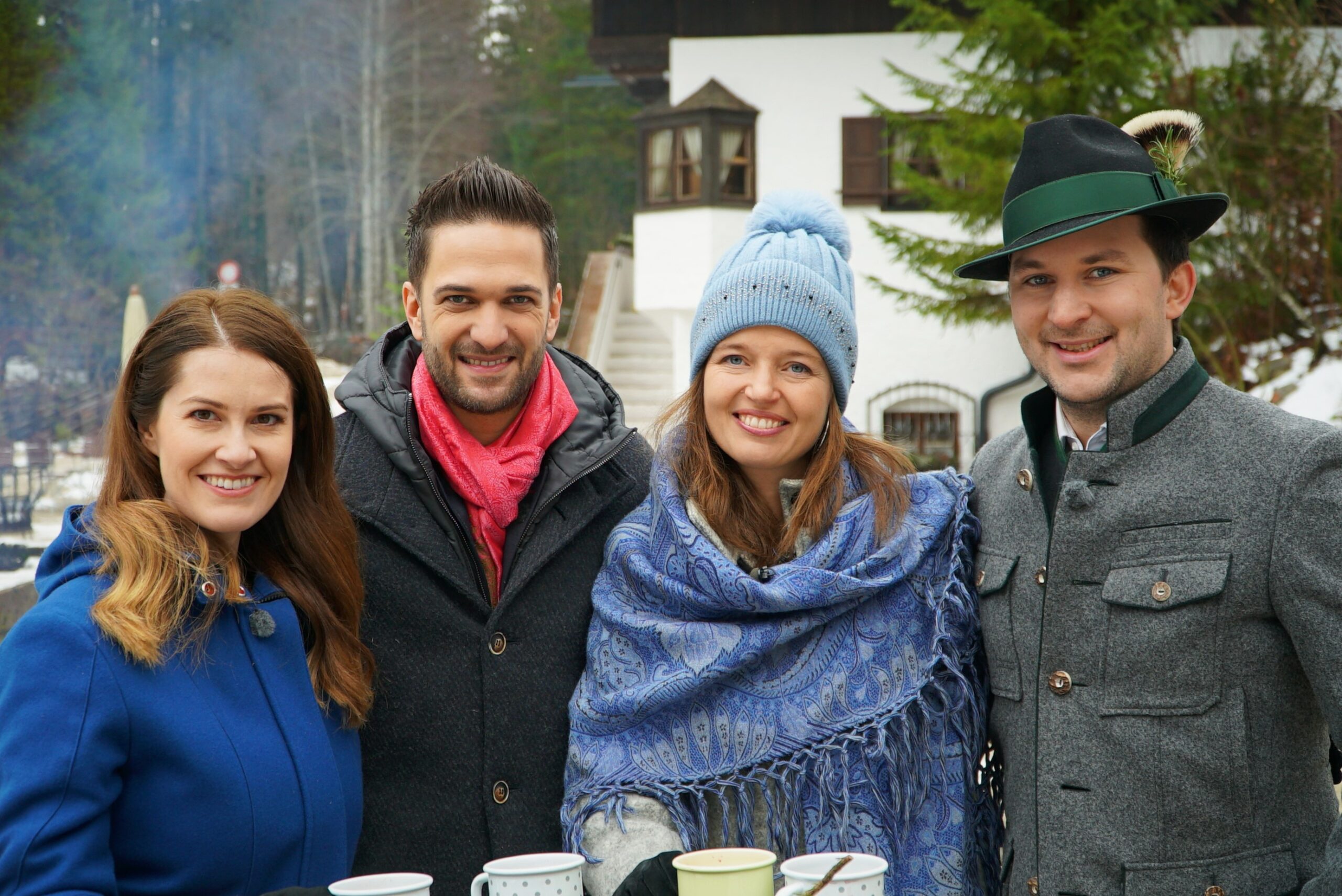 Vier lächelnde Menschen in Winterkleidung stehen mit Tassen im Freien, mit Bäumen und einem Haus im Hintergrund.