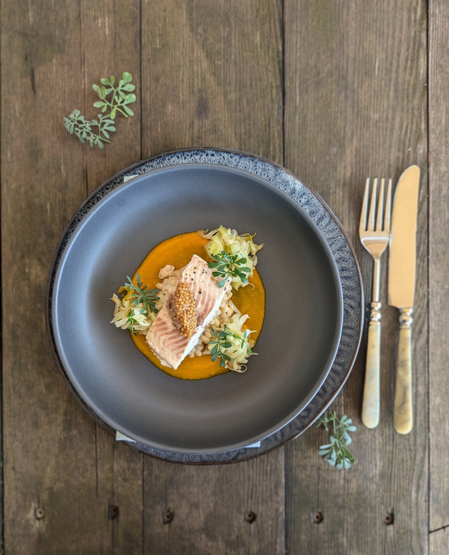 Graue Schale mit einem mit Kräutern garnierten Gourmetteller auf einem rustikalen Holztisch mit Besteck daneben.