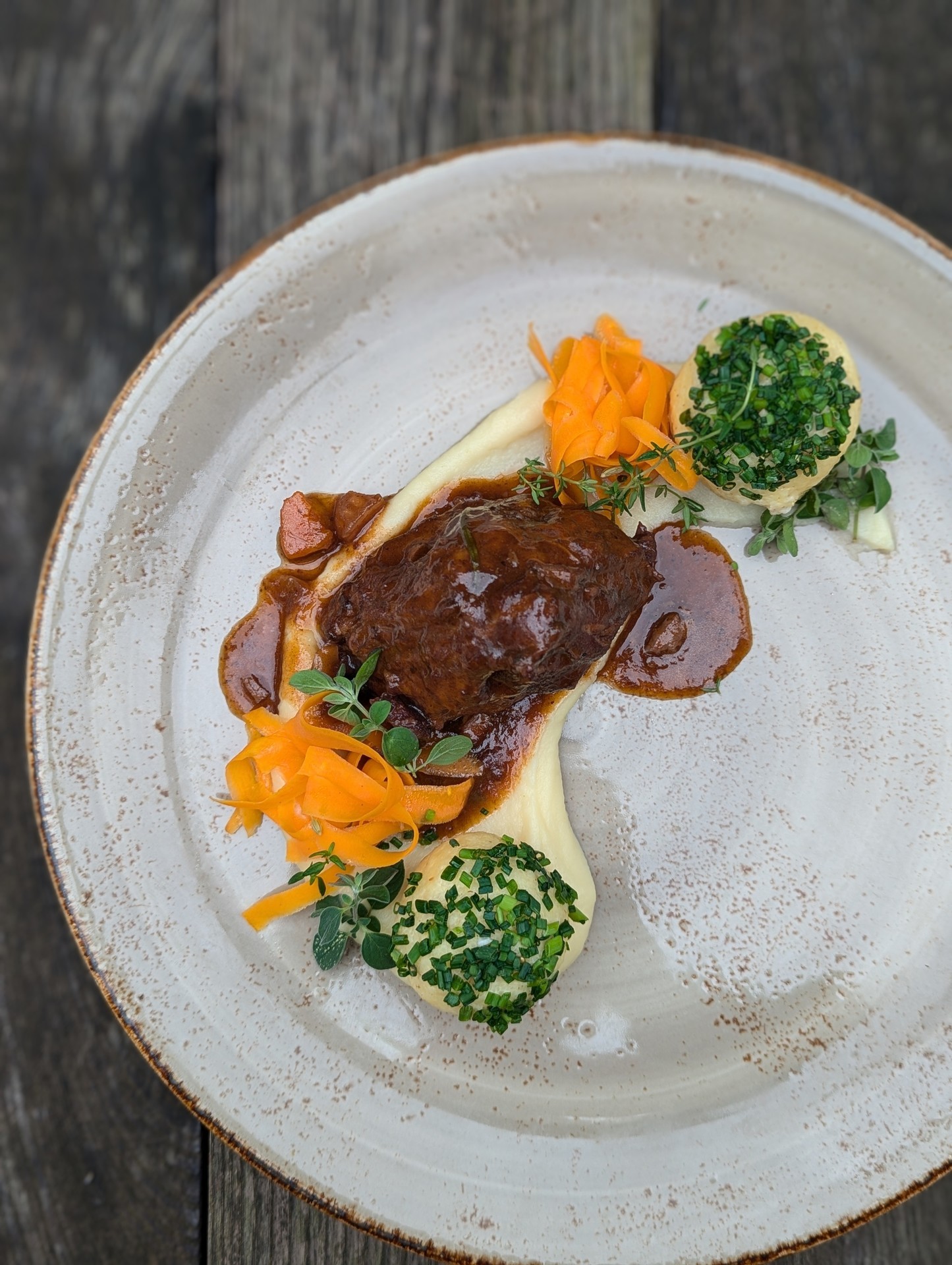 Ein Gourmetgericht mit geschmortem Fleisch, Karottenbändern, Kartoffelpüree und grüner Kräutergarnitur auf einem rustikalen Teller.