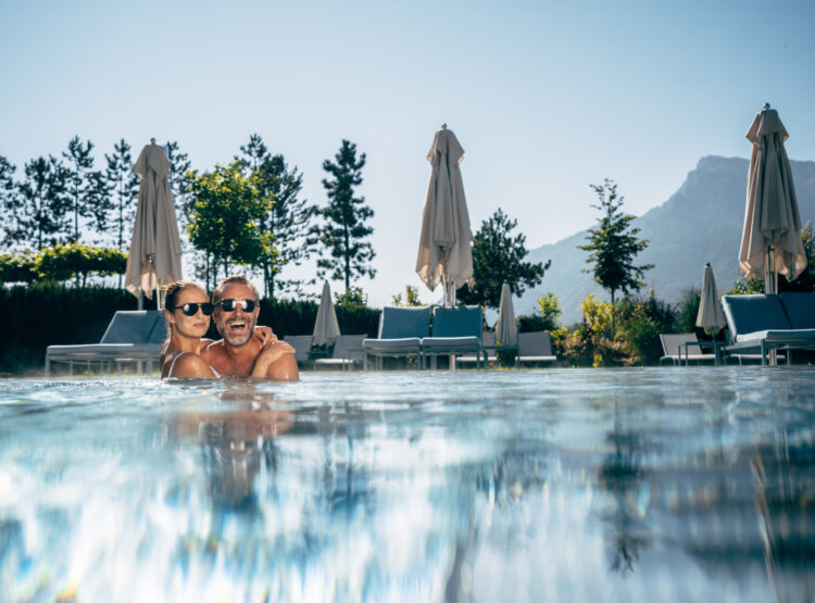 Ein lächelndes Paar mit Sonnenbrille entspannt sich am Pool des Laschenskyhofs, mit Liegestühlen und Bergen im Hintergrund.
