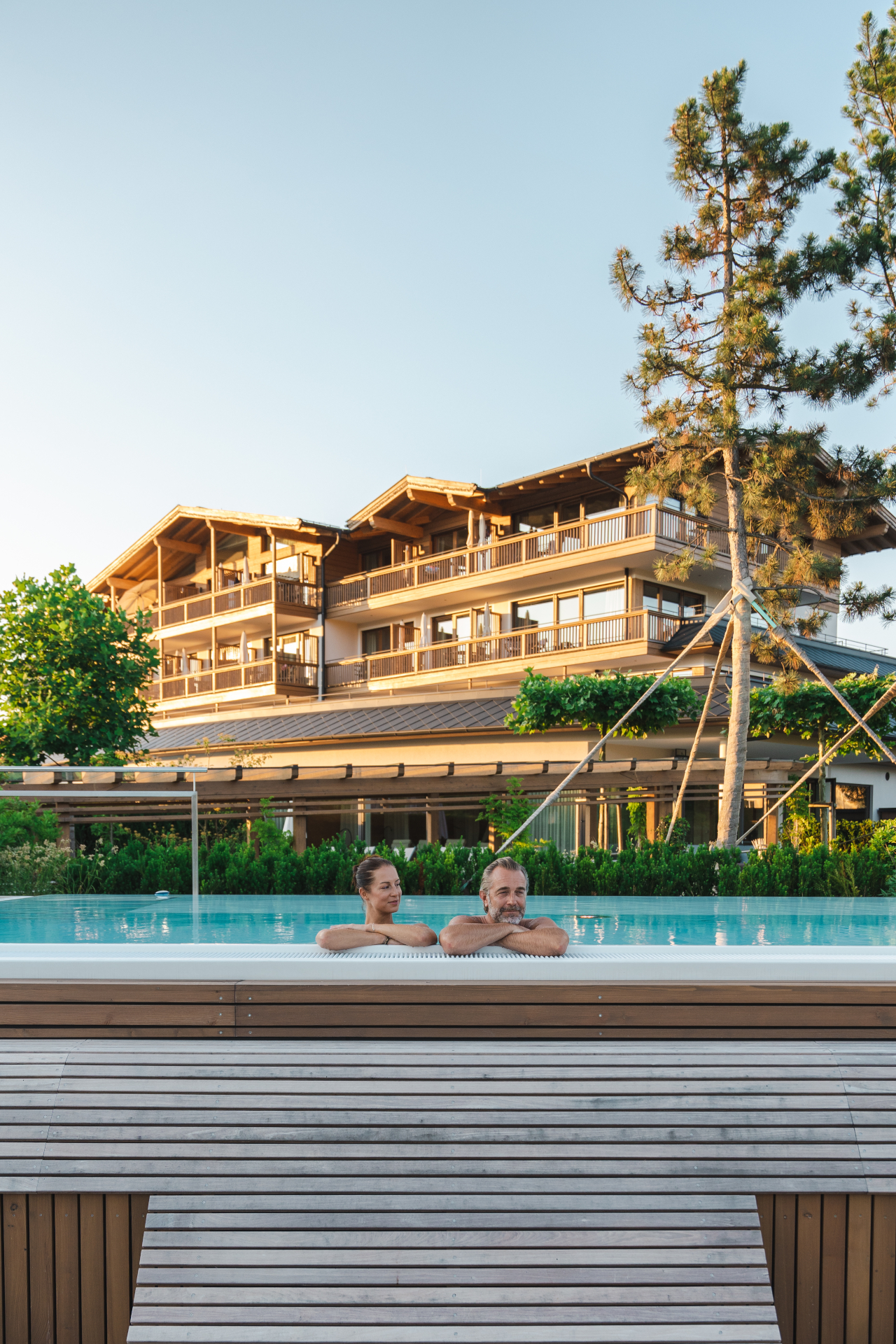 Ein l&auml;chelndes Paar entspannt sich im Au&szlig;enpool des Laschenskyhofs, der vor einem gro&szlig;en, modernen Hotelgeb&auml;ude aus Holz liegt.