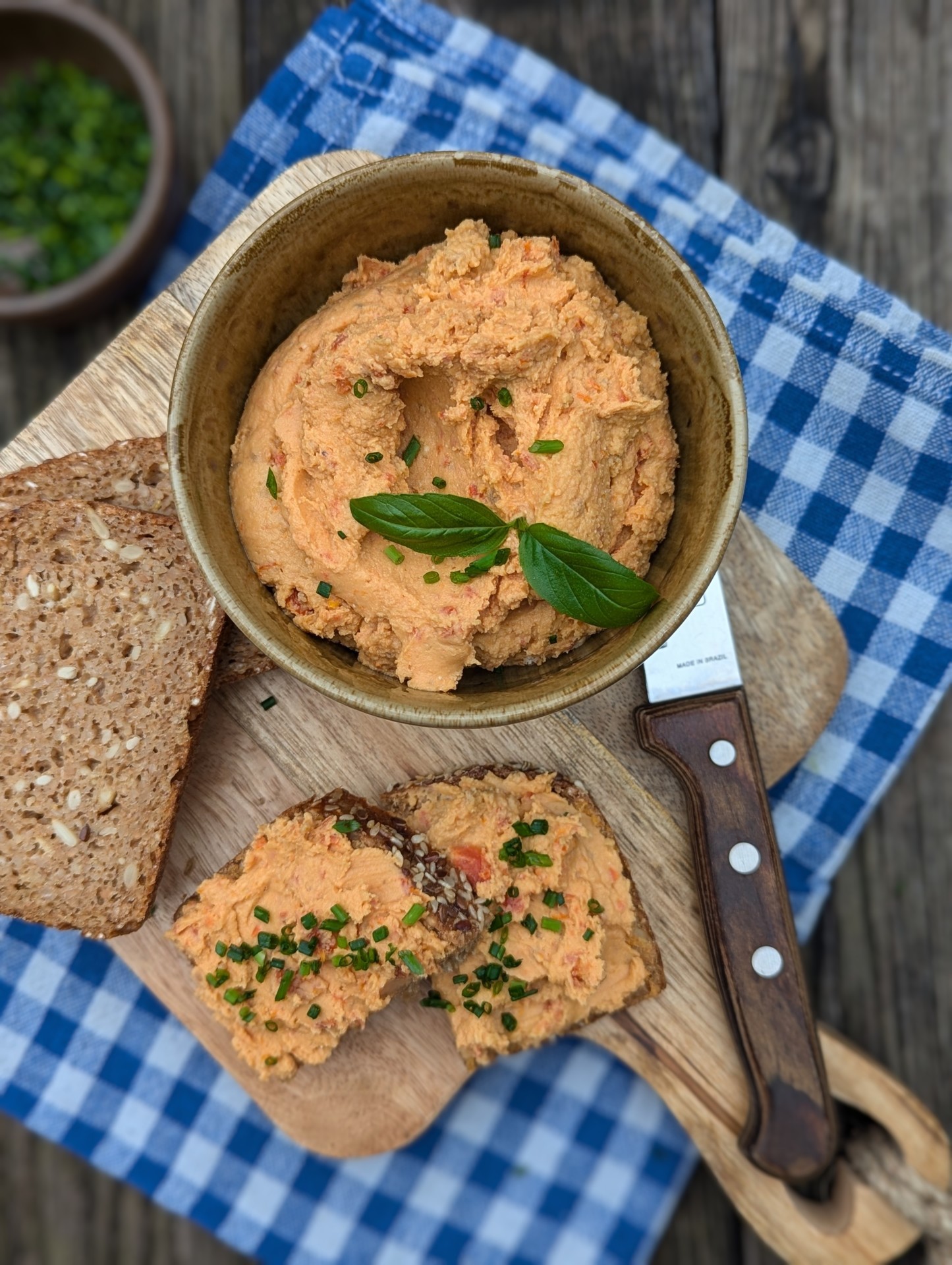 Eine Schale mit cremigem Aufstrich mit Kräutern, Brotscheiben und einem Messer auf einem blau karierten Tuch.