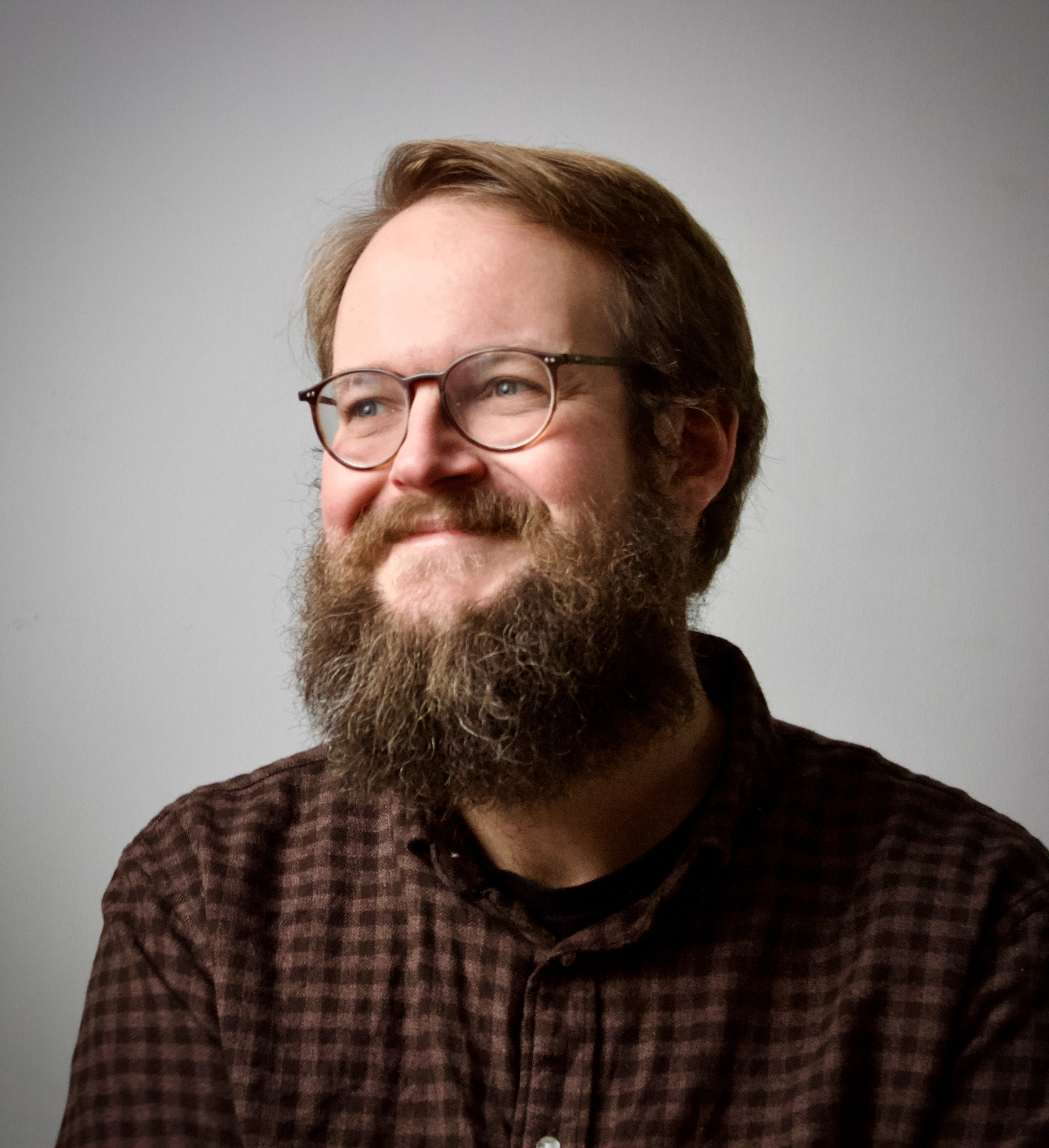 Ein b&auml;rtiger Mann mit Brille l&auml;chelt in einem karierten Hemd vor einem schlichten hellen Hintergrund.