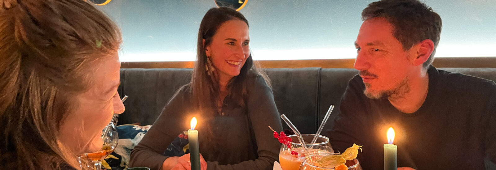 Drei Personen sitzen an einem kerzenbeleuchteten Tisch in einem Restaurant mit runden Spiegeln an der Wand, lächeln und unterhalten sich bei bunten Getränken.