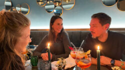 Drei Personen sitzen an einem kerzenbeleuchteten Tisch in einem Restaurant mit runden Spiegeln an der Wand, lächeln und unterhalten sich bei bunten Getränken.