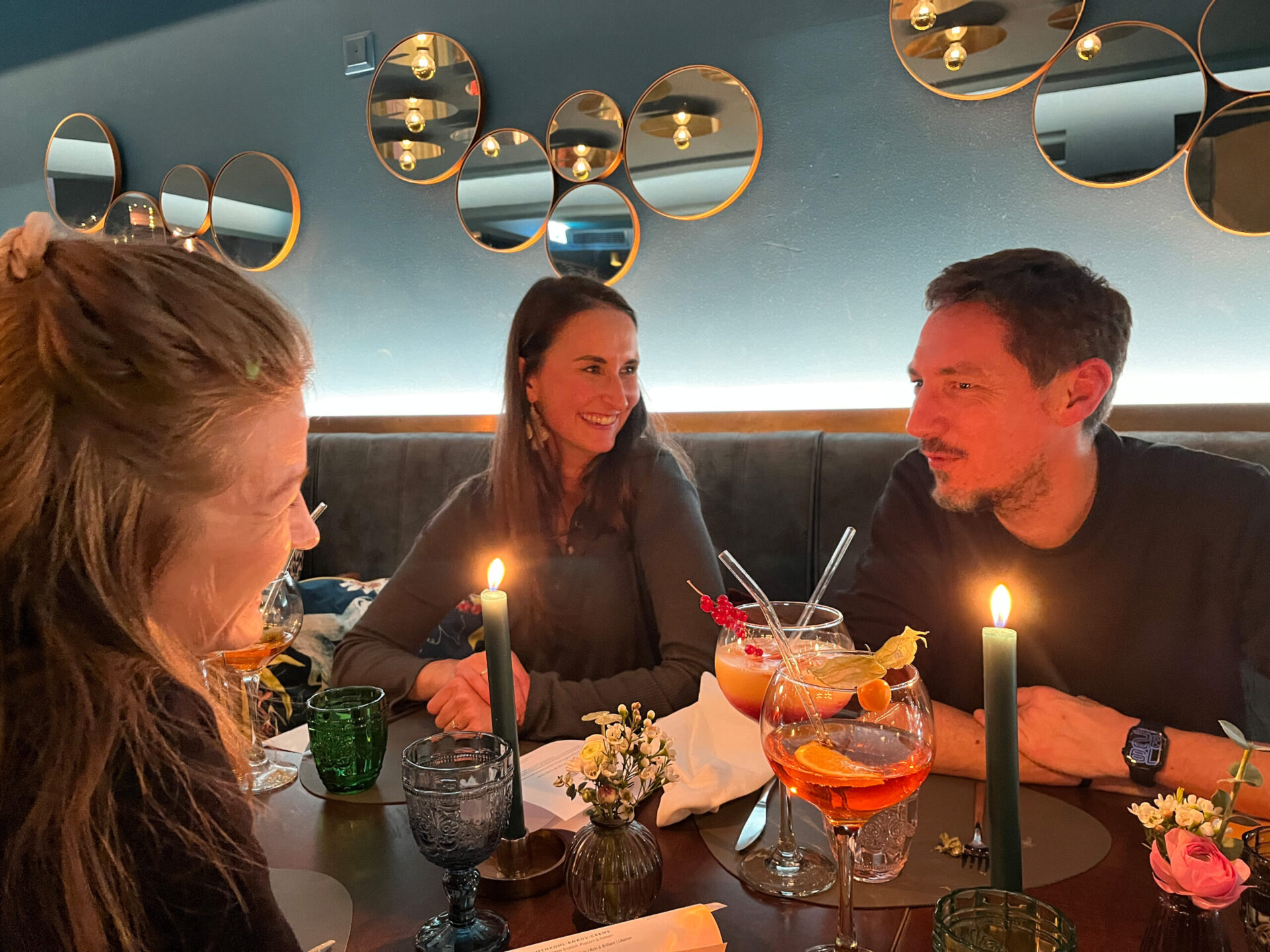 Drei Personen sitzen an einem kerzenbeleuchteten Tisch in einem Restaurant mit runden Spiegeln an der Wand, l&auml;cheln und unterhalten sich bei bunten Getr&auml;nken.