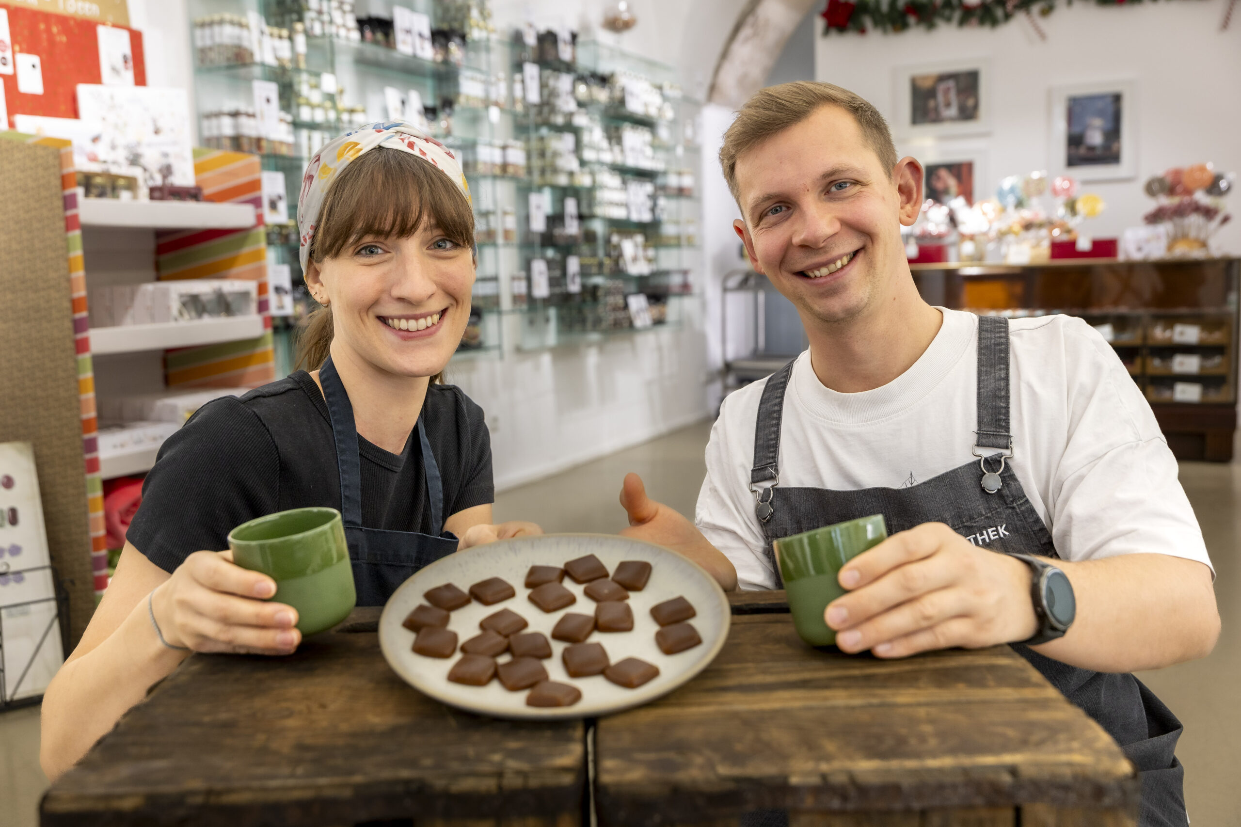 Zwei l&auml;chelnde Menschen mit gr&uuml;nen Tassen und einem Teller mit Schokolade auf einem Holztisch in einem Gesch&auml;ft.