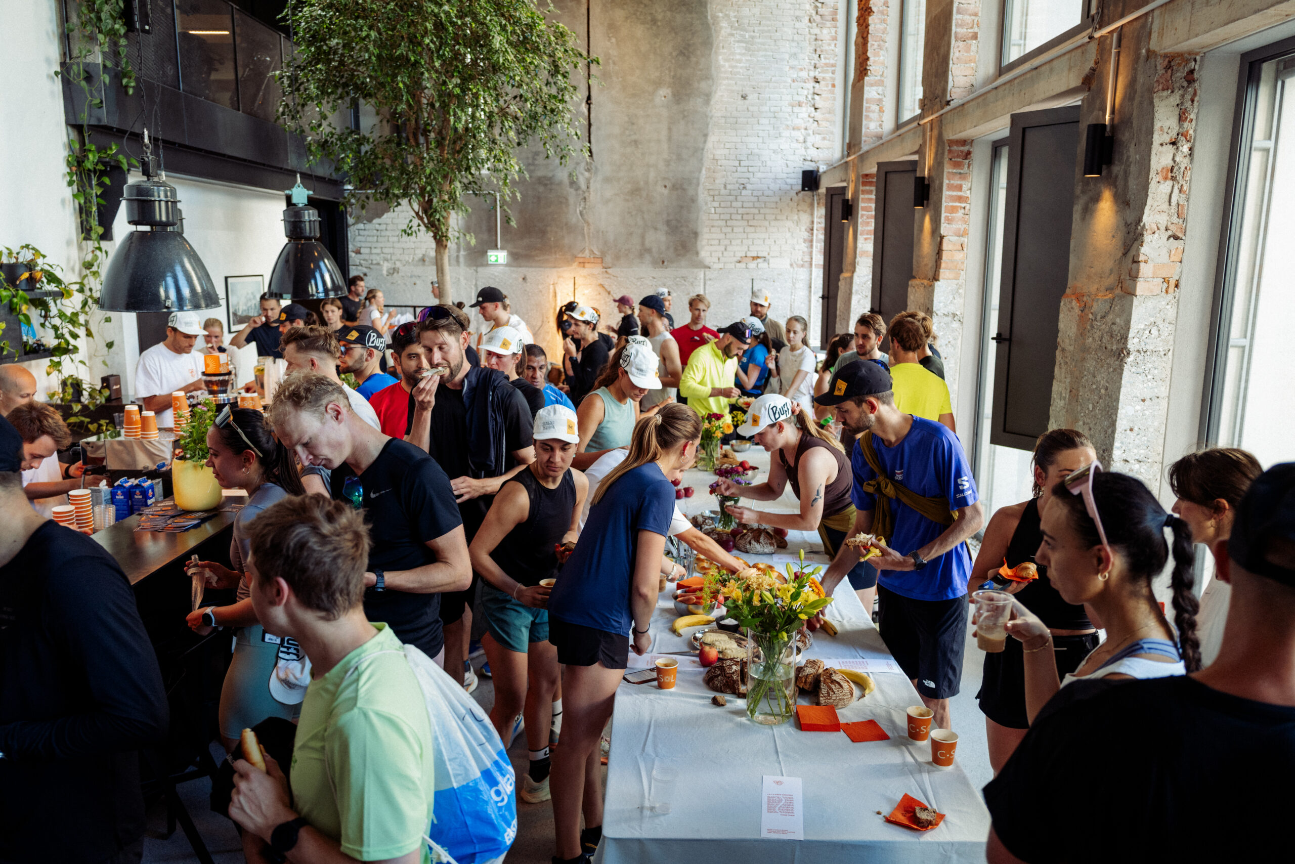 Eine gro&szlig;e Gruppe von Menschen in Sportkleidung versammelt sich um Tische mit Speisen und Getr&auml;nken in einem hellen, modernen Raum.