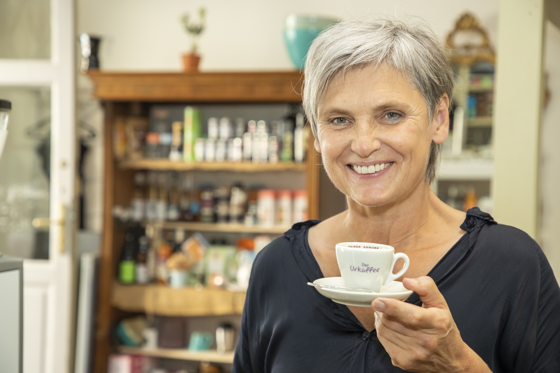 Eine l&auml;chelnde &auml;ltere Frau mit kurzen grauen Haaren h&auml;lt eine Kaffeetasse in einer gem&uuml;tlichen K&uuml;che oder in einem Caf&eacute;.