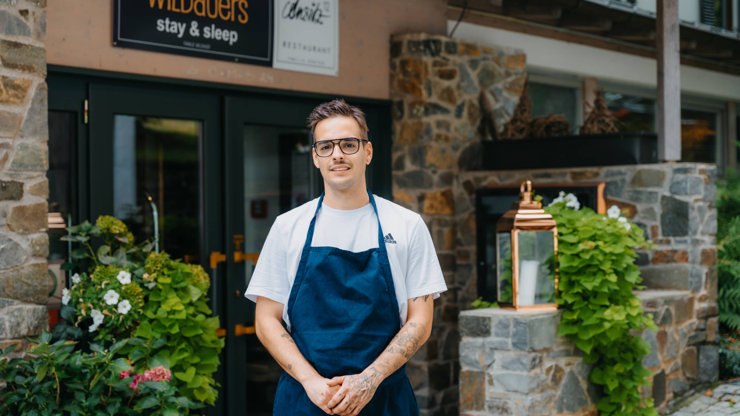 Eine Person mit Brille und blauer Schürze steht lächelnd vor einem Steinhaus mit Pflanzen.