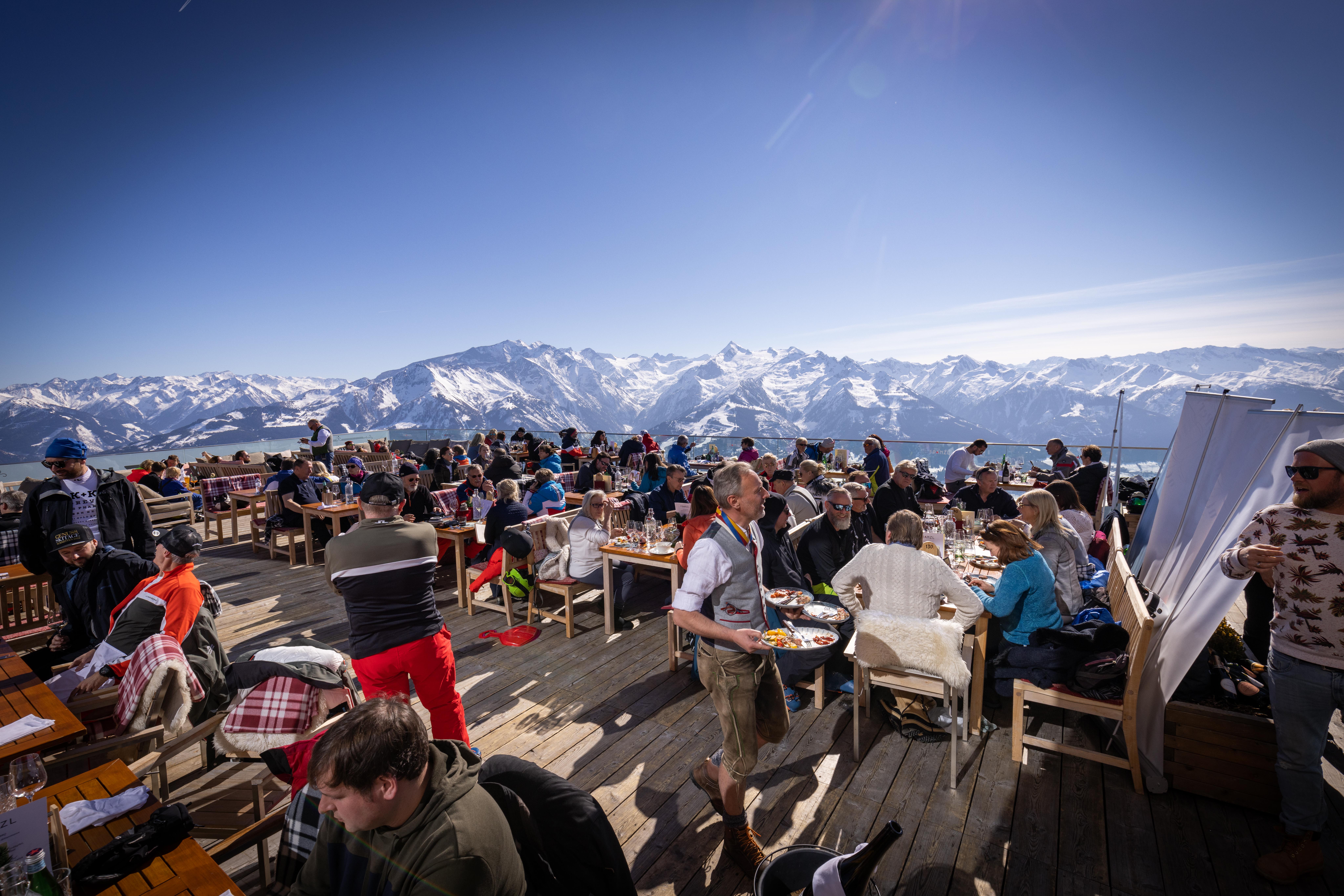 Menschen essen im Freien auf einer Holzterrasse mit schneebedeckten Bergen im Hintergrund unter einem klaren blauen Himmel.