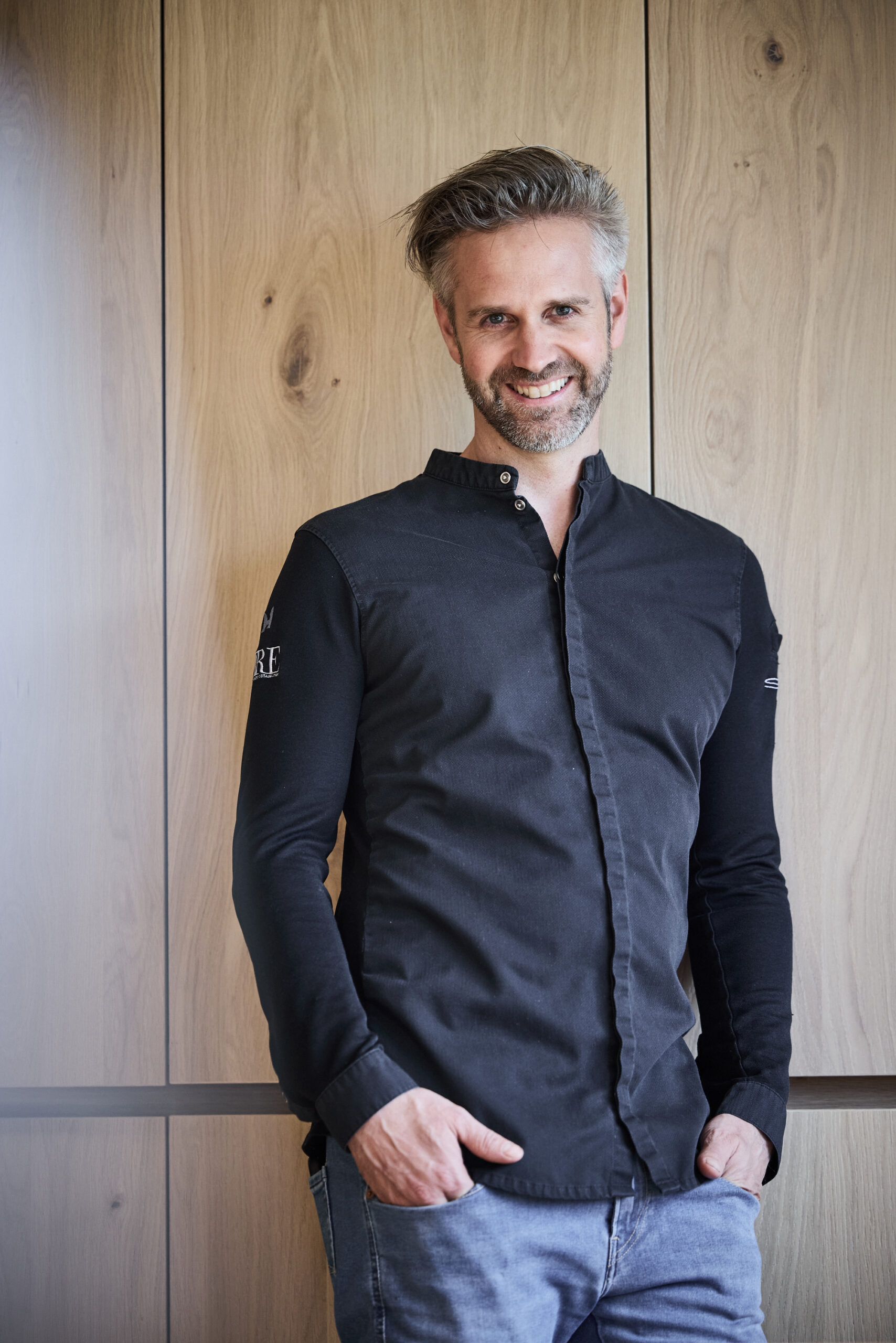 Mann mit grauem Haar und Bart, der ein schwarzes Hemd und Jeans tr&auml;gt, l&auml;chelt vor einer Holzwand.