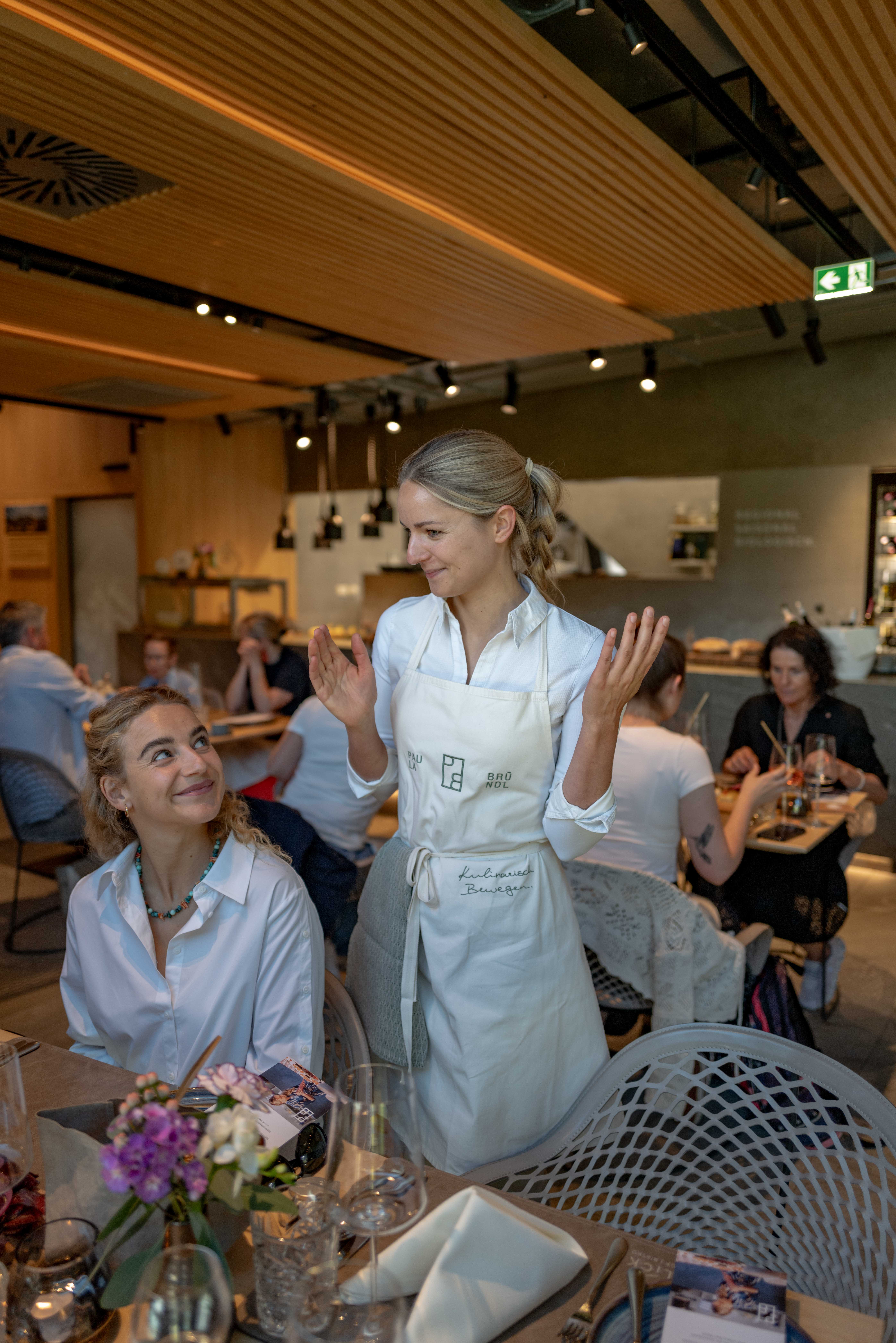 Eine Kellnerin l&auml;chelt und gestikuliert, w&auml;hrend sie mit einer sitzenden Frau in einem belebten, modernen Restaurant spricht.
