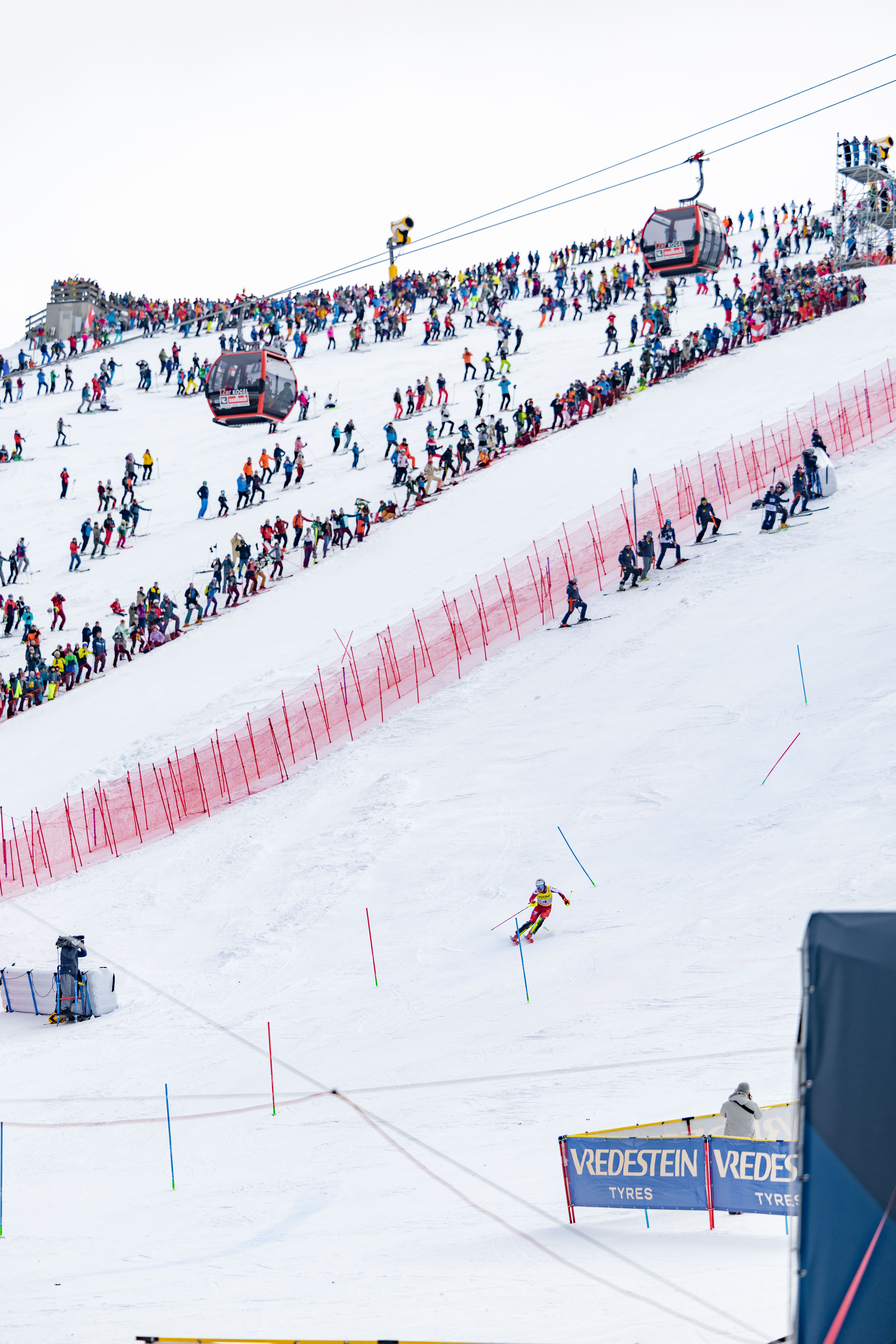 Ein Skifahrer fährt bergab, während die Zuschauer von einer verschneiten Piste aus zusehen, über der Seilbahnen und Zäune die Strecke säumen.