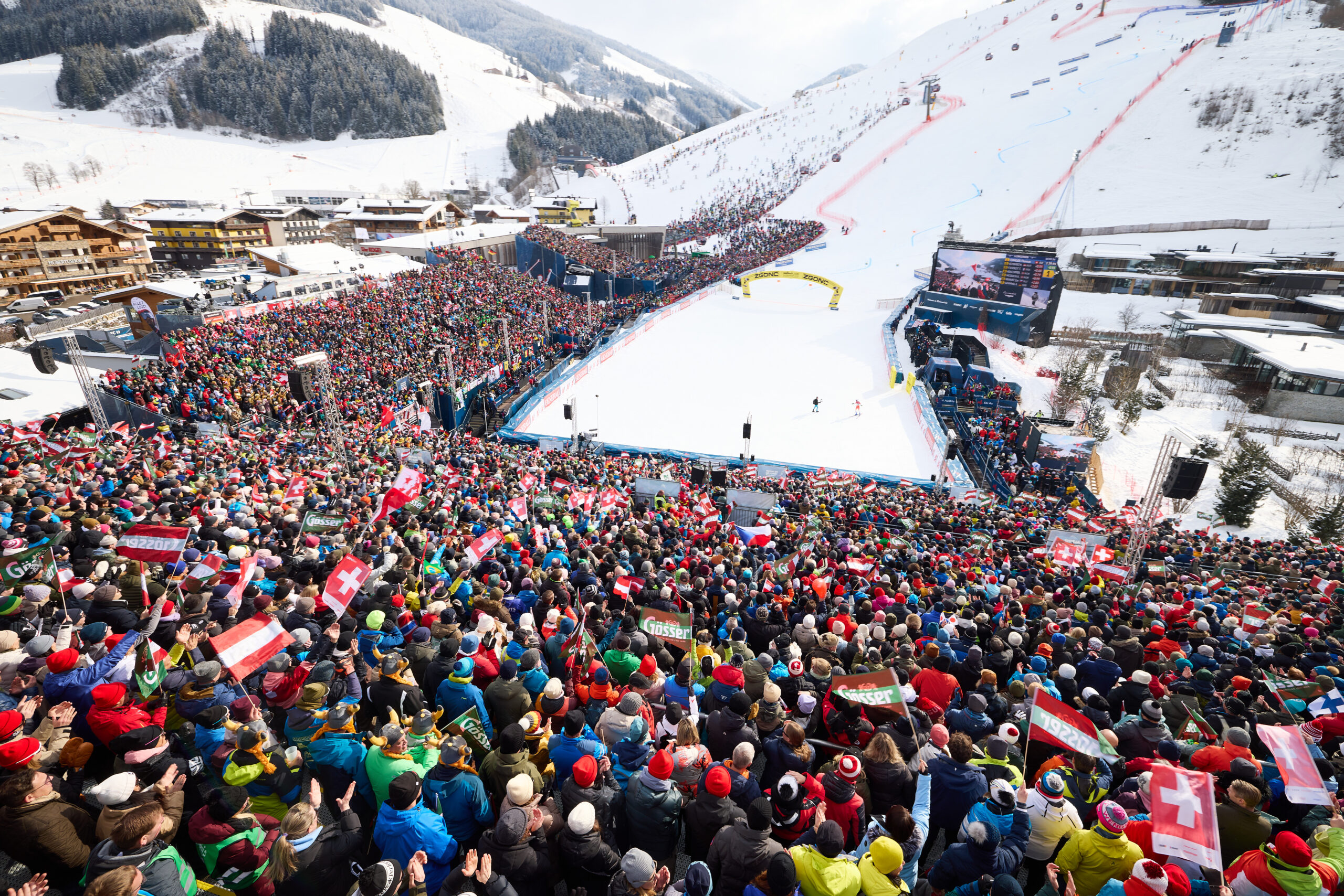 Große jubelnde Menschenmenge bei einer verschneiten Skiveranstaltung mit Bergen, Gebäuden und einer Skipiste im Hintergrund.