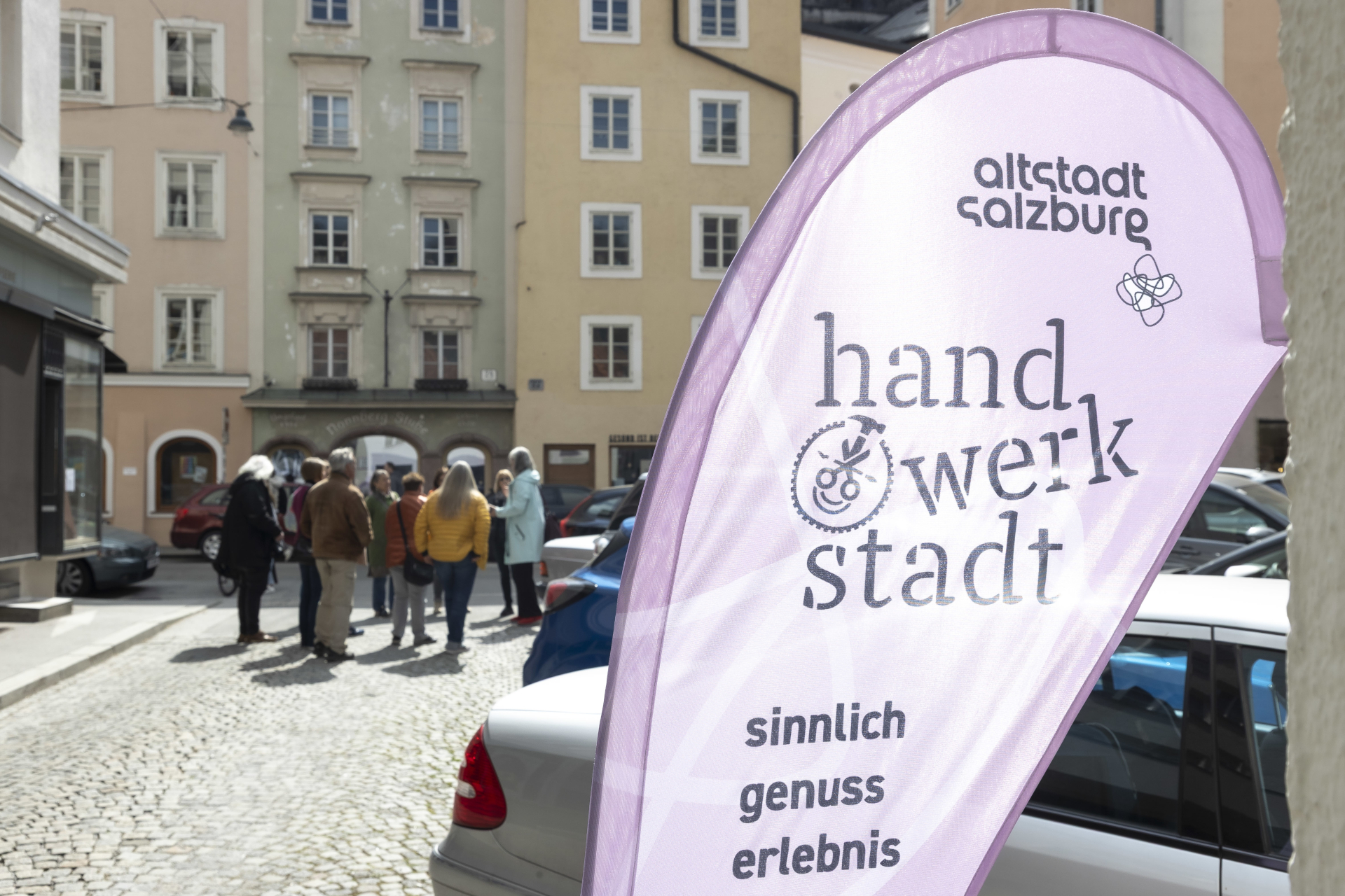 Lila Banner mit der Aufschrift "handwerkstatt" auf Deutsch, mit Geb&auml;uden und einer Gruppe von Menschen im Hintergrund.