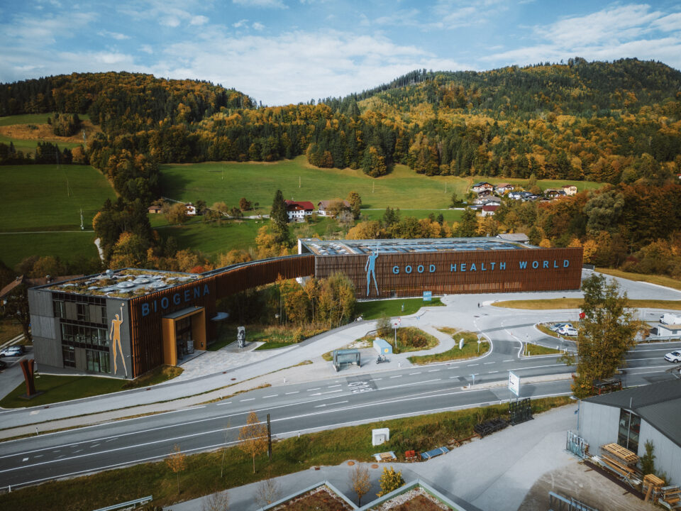 Ein modernes Gebäude mit der Aufschrift Biogena Good Health World, umgeben von Bäumen und Hügeln im Herbst.