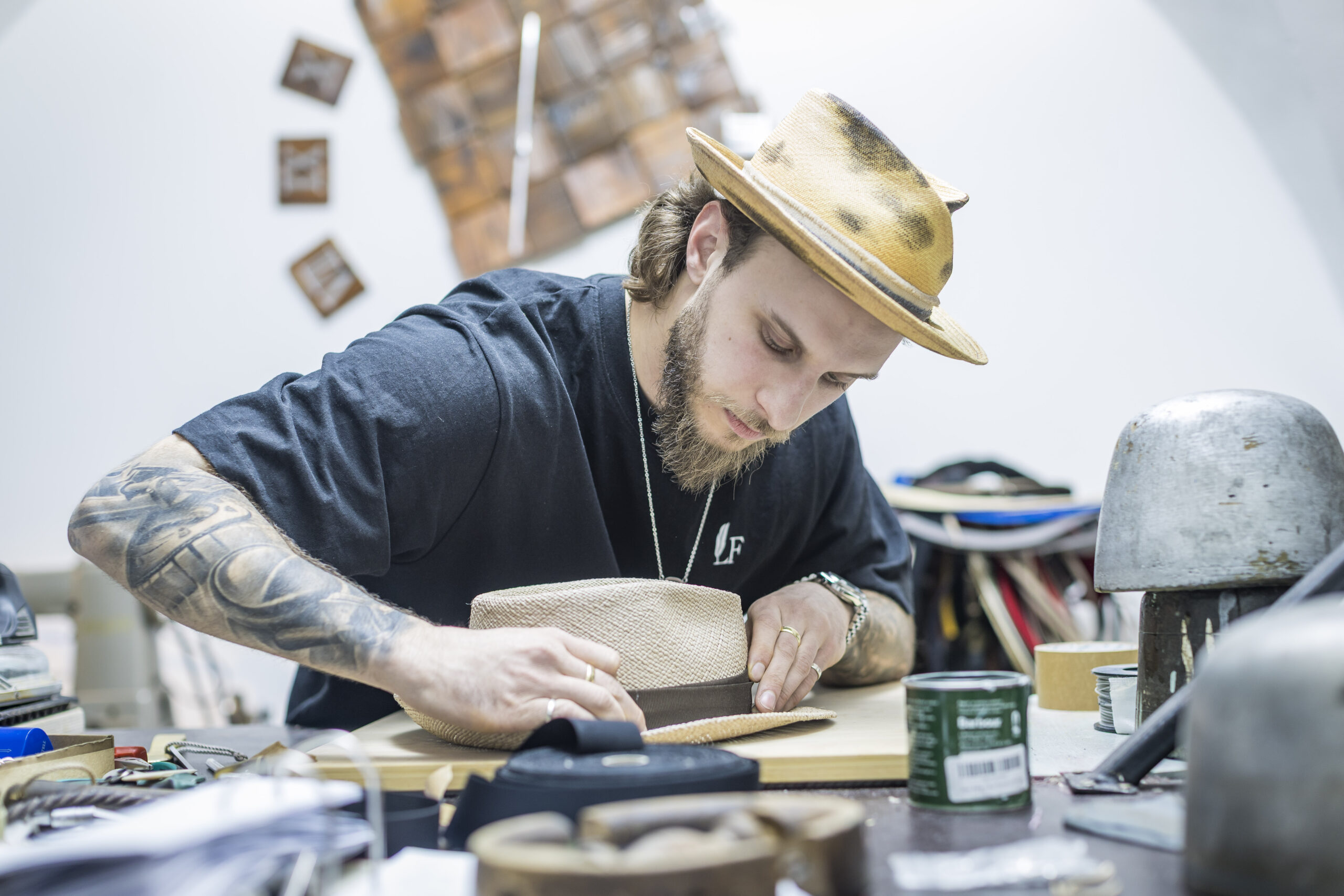 Ein Mann mit T&auml;towierungen formt einen Hut an einer &uuml;berf&uuml;llten Werkbank in einer Handwerkerwerkstatt.