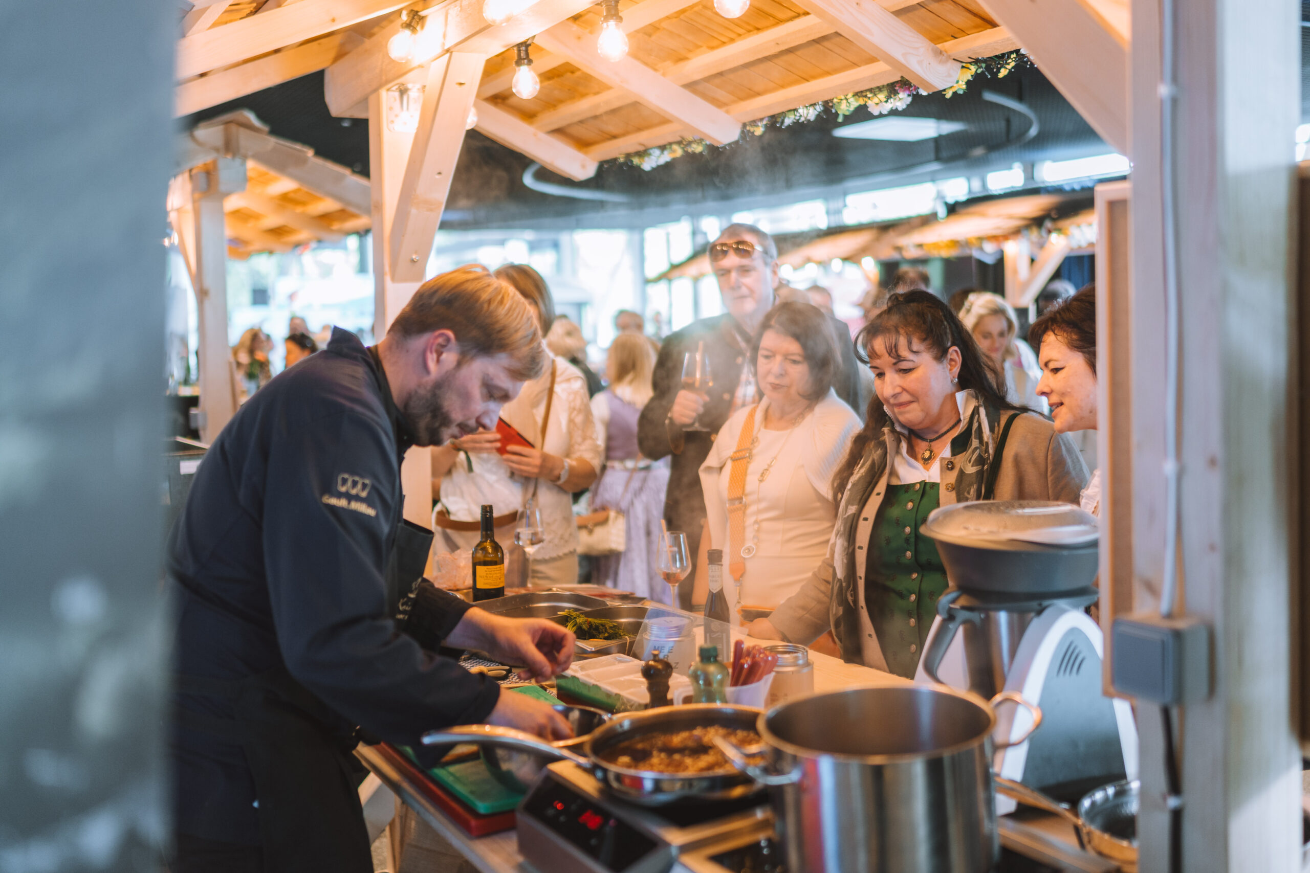 Ein Koch bereitet an einem Marktstand Lebensmittel zu, während eine Gruppe auf dem mundART Festival aufmerksam zusieht.