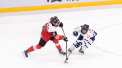 Zwei Eishockeyspieler in Aktion, einer in Rot und Schwarz, einer in Weiß und Blau, kämpfen um den Puck auf dem Eis.