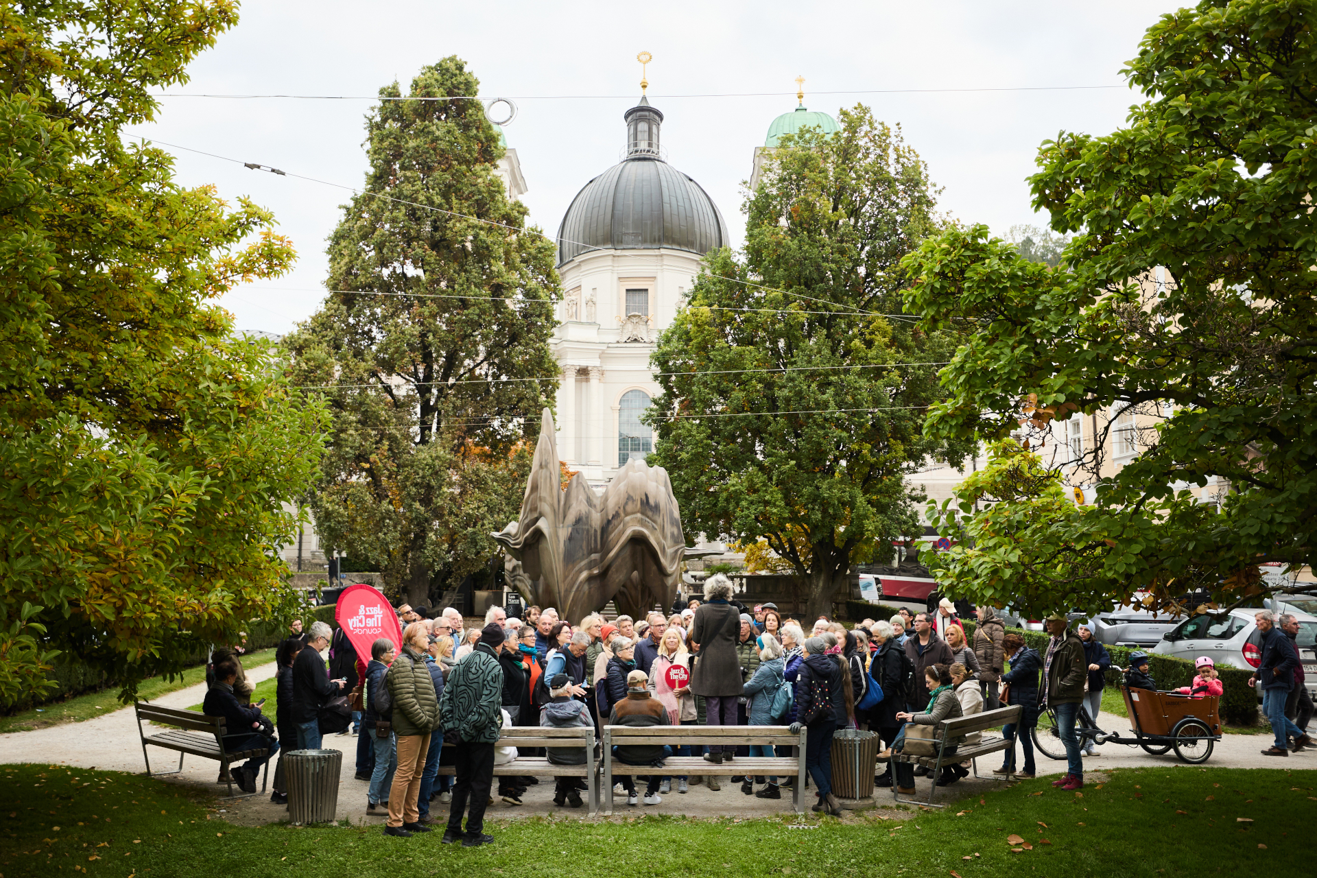 Ein Reiseleiter spricht zu einer Gruppe von Menschen in einem Park, im Hintergrund ist eine Kuppelkirche zu sehen.