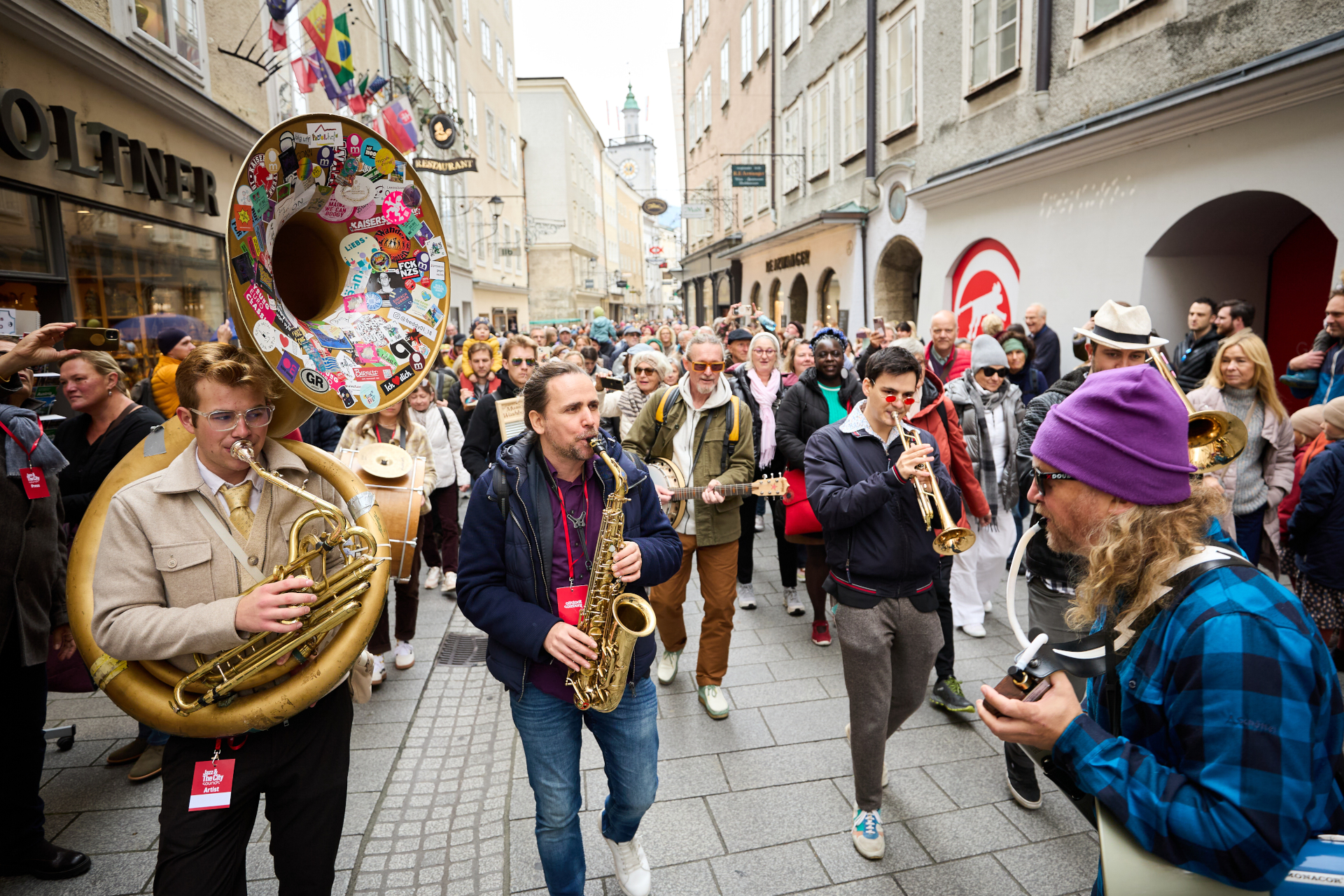 Eine Straßenkapelle spielt auf Blechblasinstrumenten, während eine Menschenmenge zusieht und eine Stadtstraße entlanggeht.