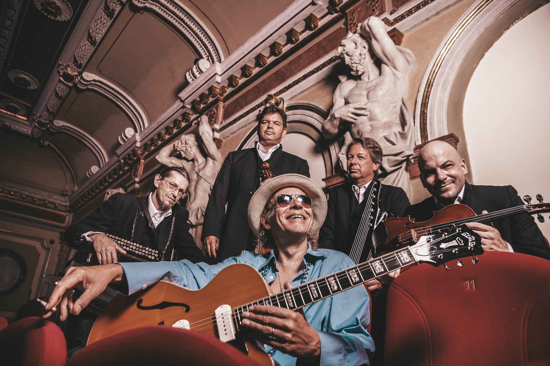 Fünf Musiker sitzen in kunstvoll verzierten Theatersesseln; einer sitzt vorne, hält eine Gitarre in der Hand und lächelt, hinter ihnen stehen Statuen.