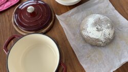 Runder Teig auf Pergament neben einem leeren roten Dutch Oven und Keramikschüsseln auf einem Holztisch.