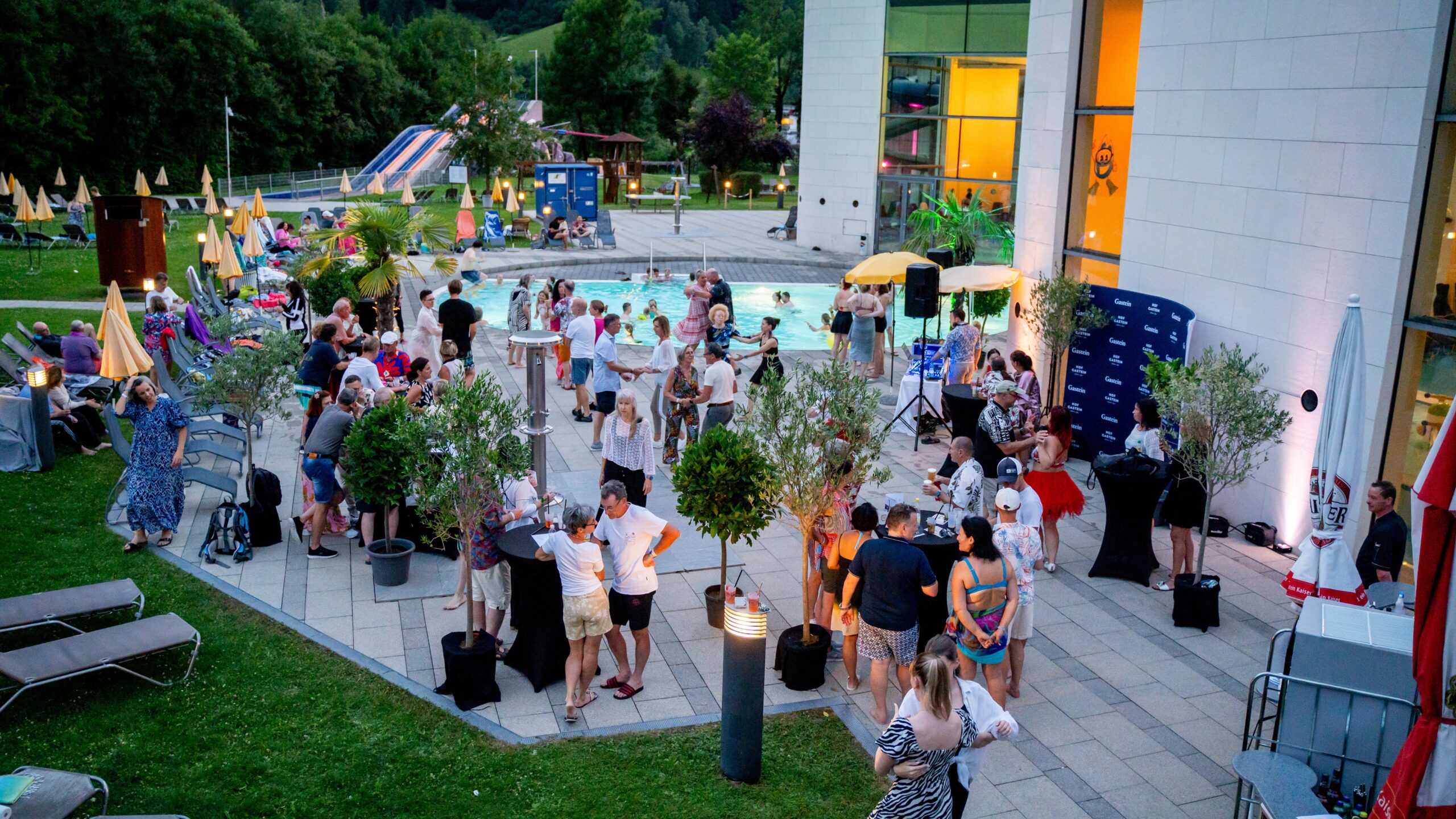 Menschen versammeln sich bei einer Veranstaltung im Freien um einen Pool mit Liegestühlen, Sonnenschirmen und Bäumen in der Nähe.