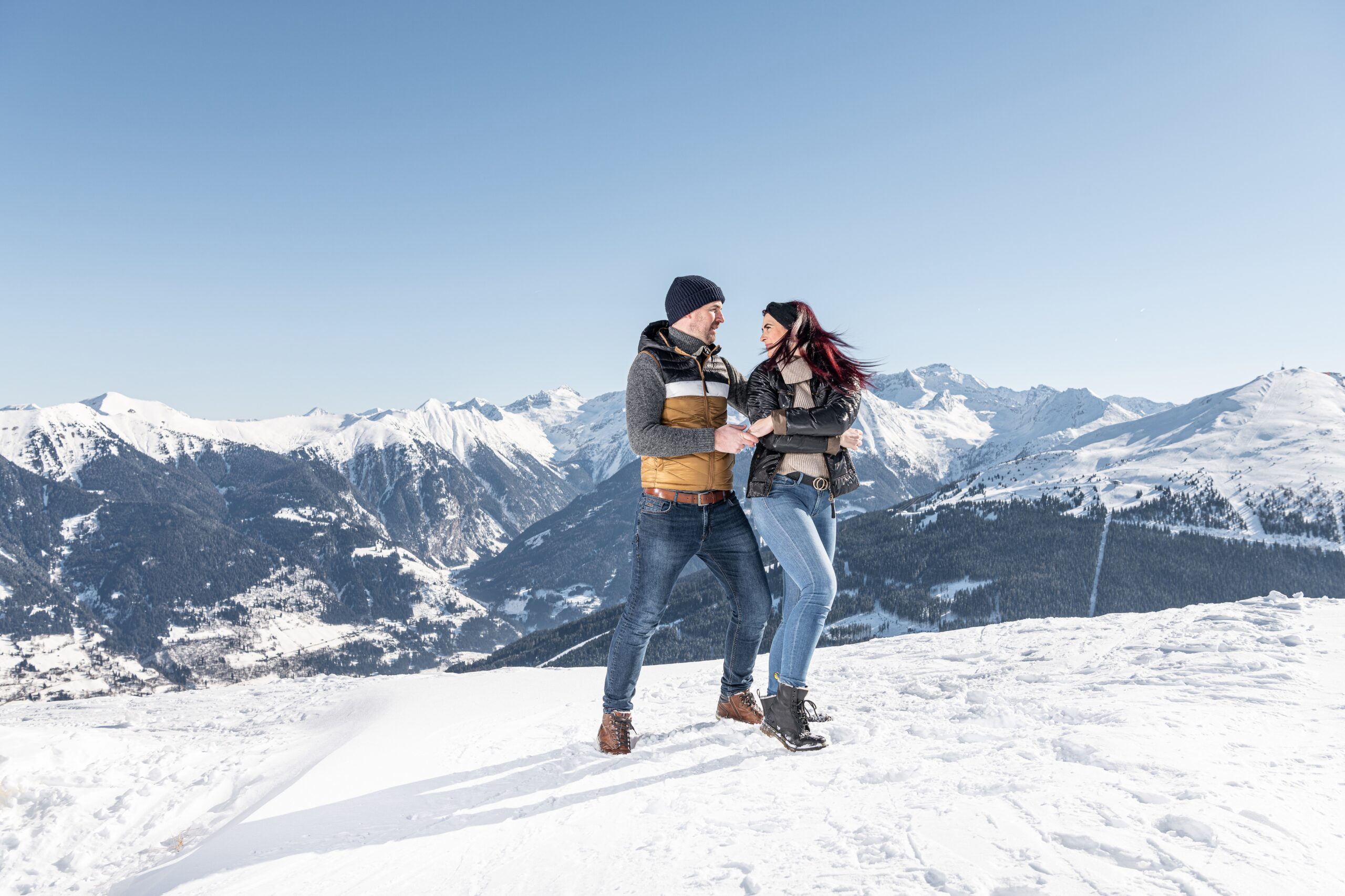 Ein Paar lächelt und hält sich an den Händen, während es auf einem verschneiten Berg mit malerischen Gipfeln im Hintergrund steht.