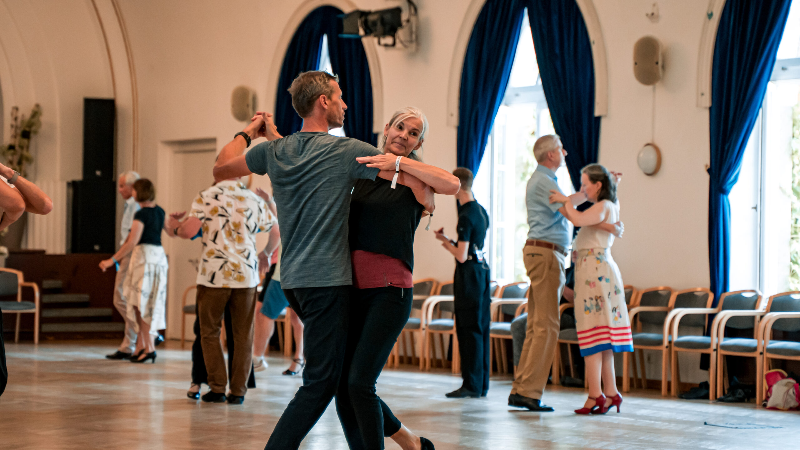 Paare tanzen in einem hellen Raum mit großen Fenstern und blauen Vorhängen.