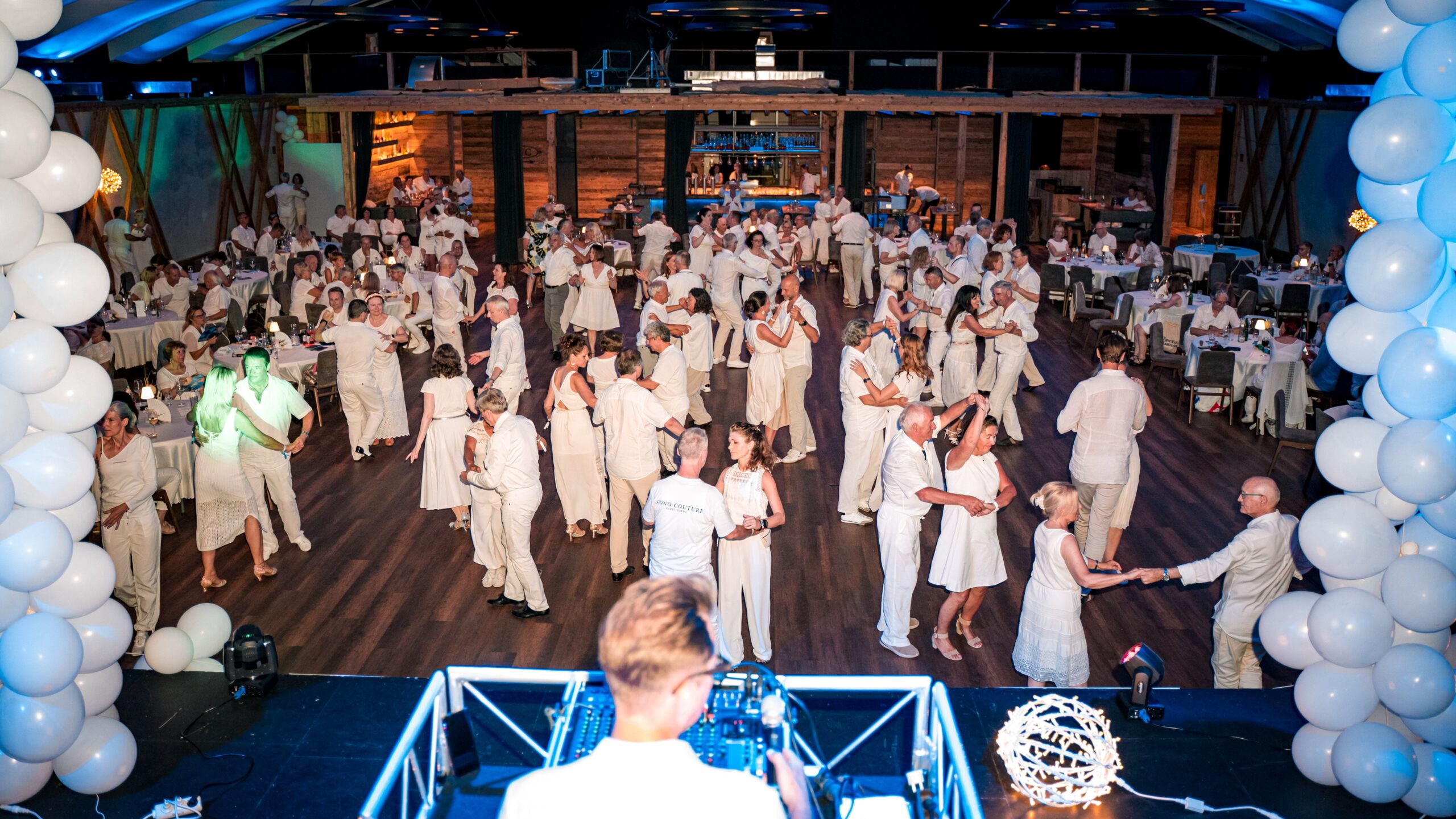 Eine große Gruppe von Menschen in weißen Kleidern tanzt auf einer lebhaften Indoor-Party mit weißen Luftballon-Dekorationen.