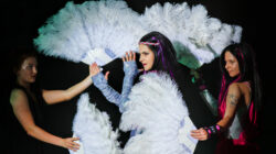 Drei Frauen in dunklen Kostümen halten große weiße Federfächer und treten vor einem schwarzen Hintergrund auf.