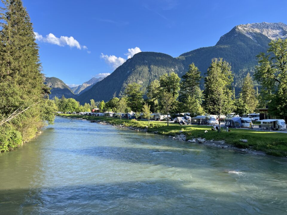 Ein Fluss fließt an einem Campingplatz mit Wohnmobilen und Zelten vorbei, umgeben von Bergen und Bäumen unter einem blauen Himmel.