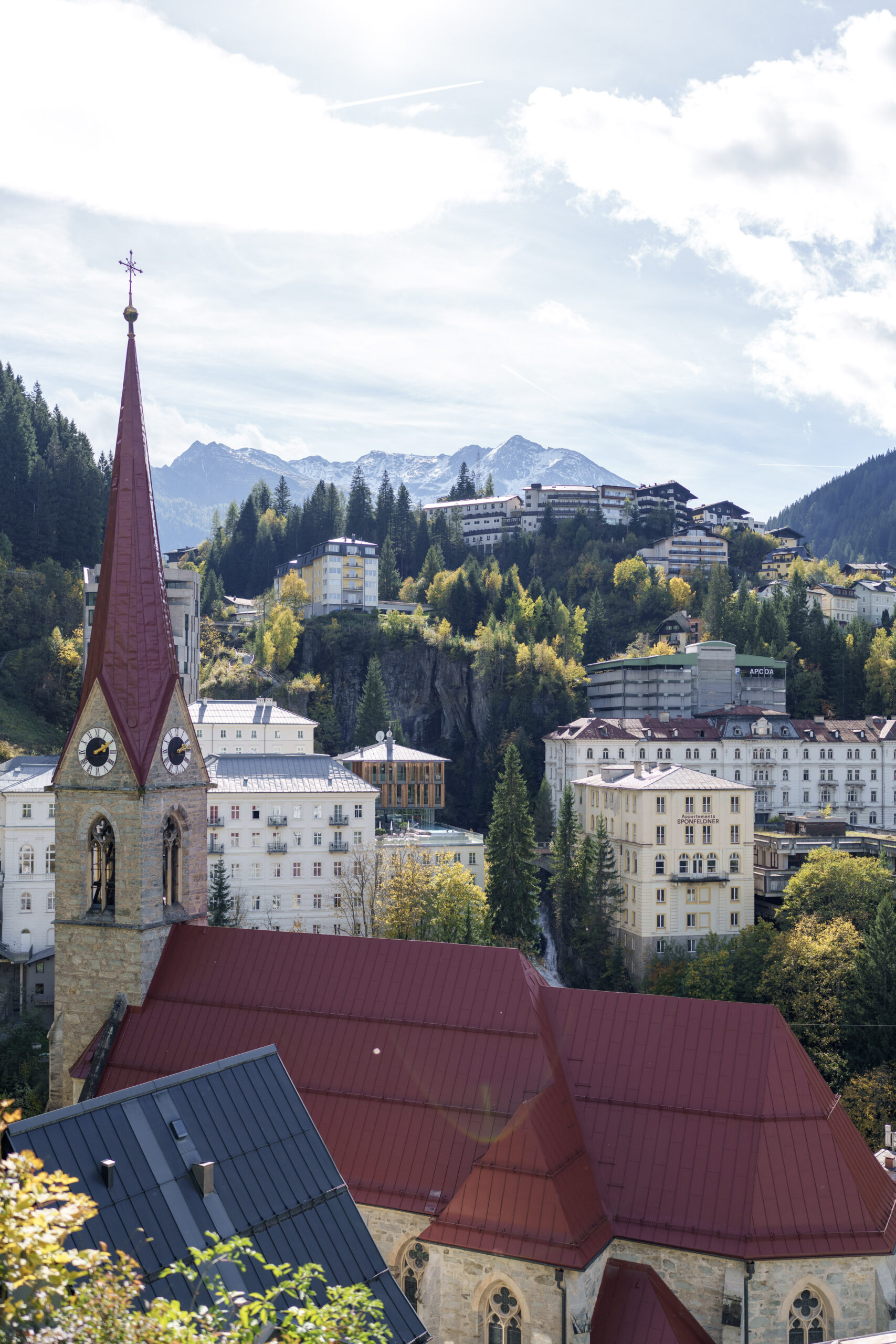 Vor einem teilweise bew&ouml;lkten Himmel erhebt sich eine Kirche mit rotem Dach und Turmspitze vor den Bergen und den am Hang gelegenen Geb&auml;uden.