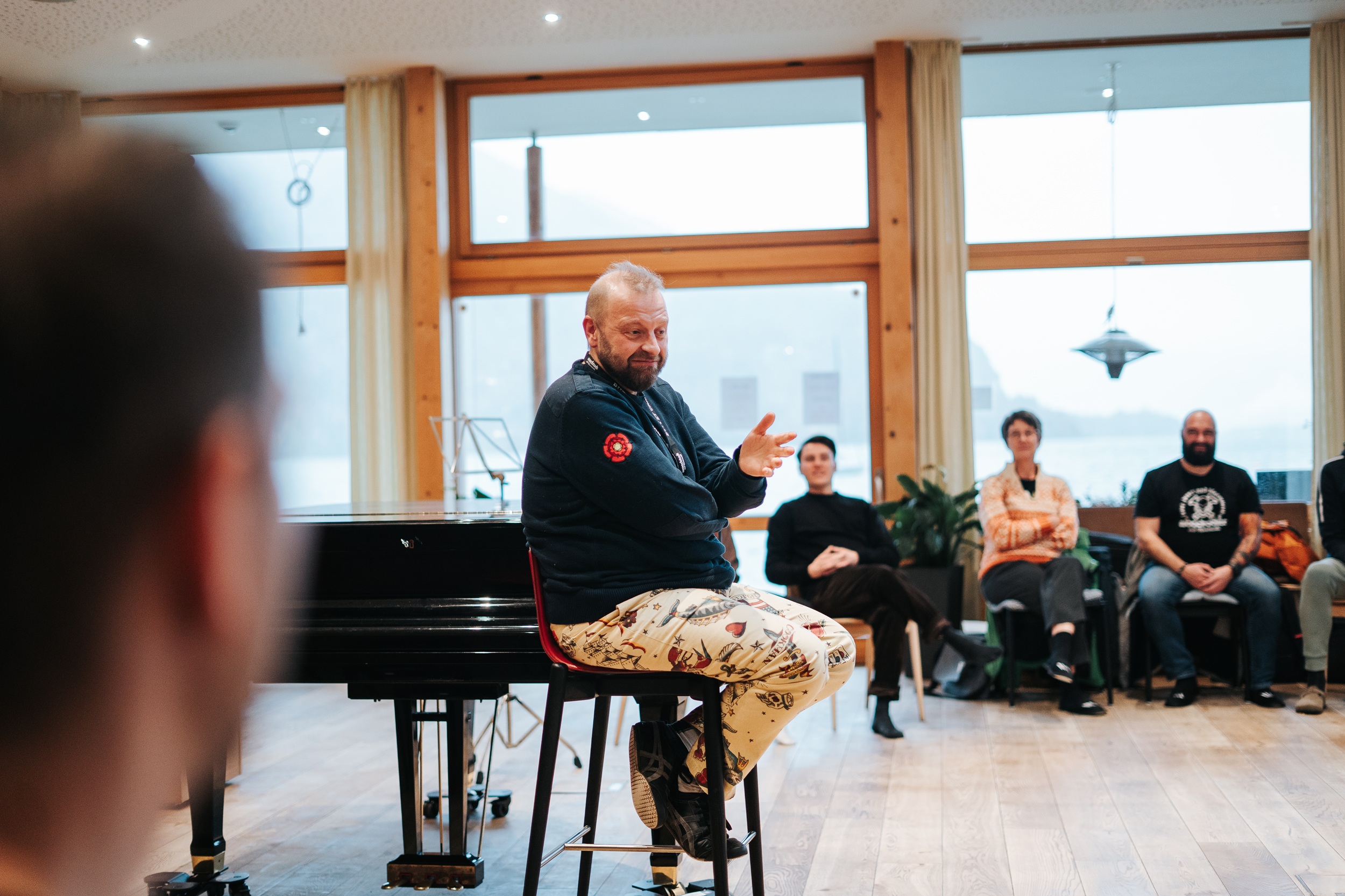 Ein Mann sitzt auf einem Hocker und spricht in der Woodstock Academy in einem hellen Raum mit gro&szlig;en Fenstern zu einer sitzenden Gruppe.