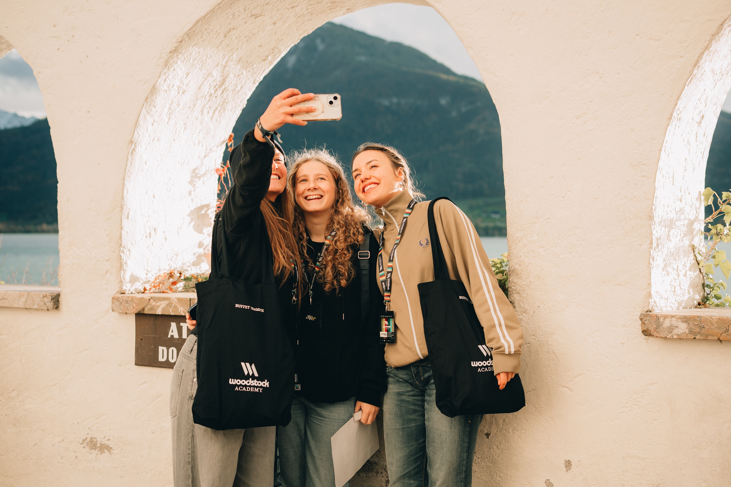 Drei junge Frauen in Sweatshirts der Woodstock Academy machen ein Selfie vor einer malerischen Berg- und Seelandschaft.