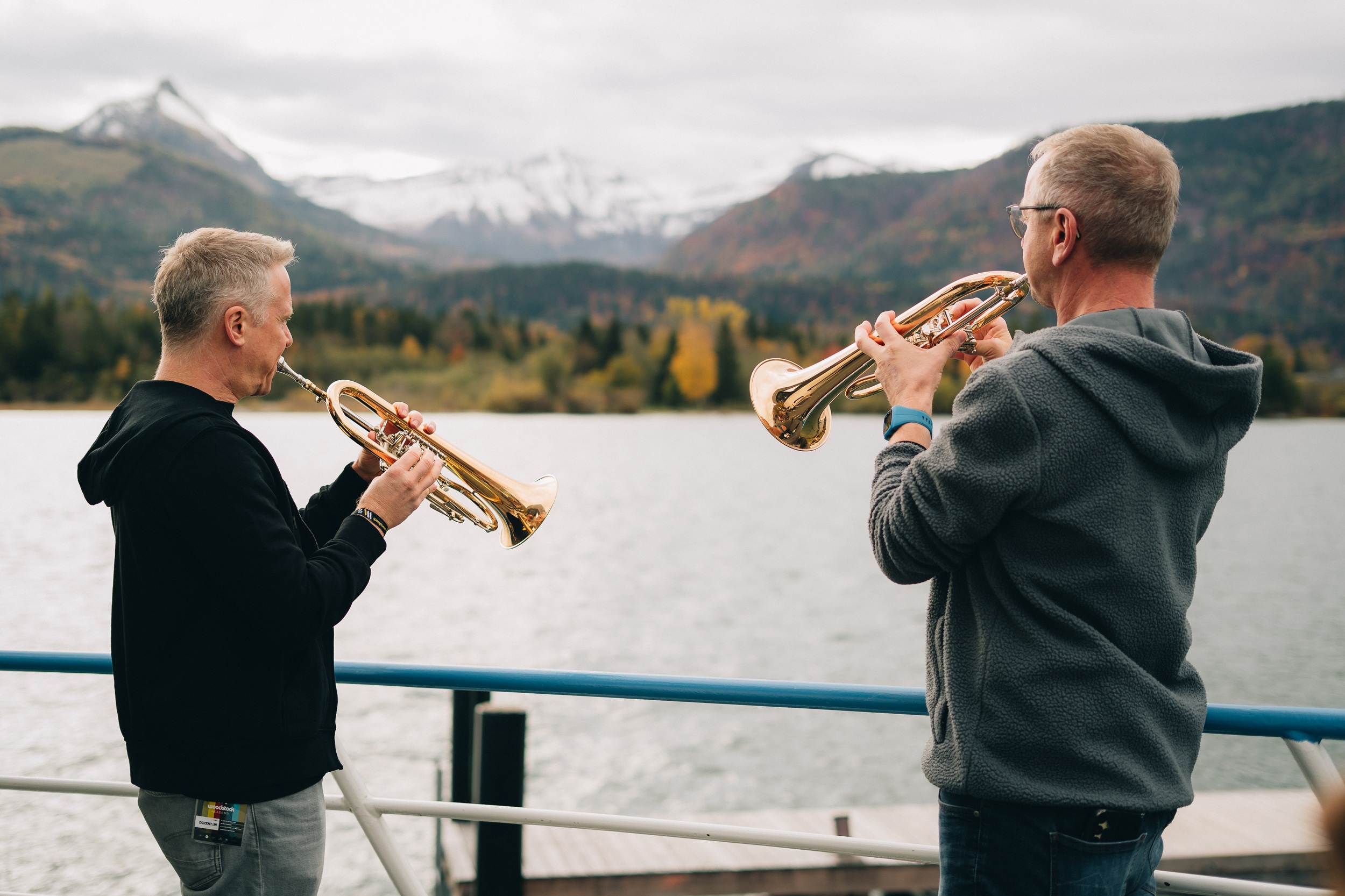 Zwei Sch&uuml;ler der Woodstock Academy spielen Blechblasinstrumente an einem See mit Bergen und B&auml;umen im Hintergrund.