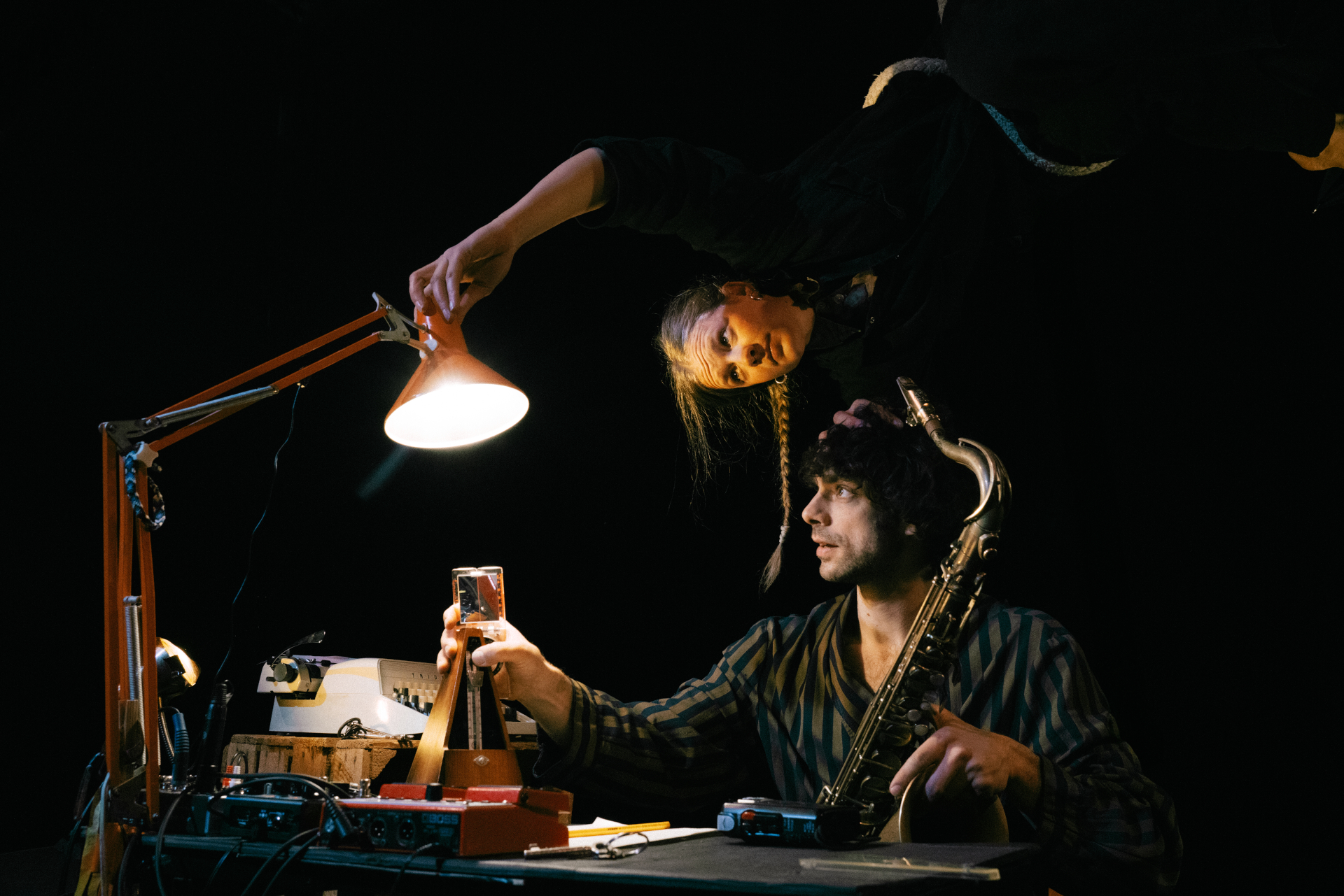 Eine Frau richtet eine Lampe auf einen Musiker mit einem Saxophon, der an einem mit Instrumenten überfüllten Schreibtisch sitzt.