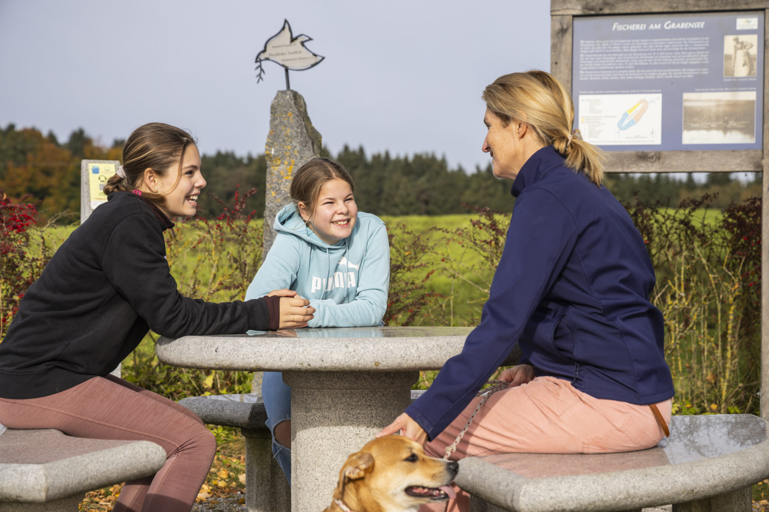 Drei Frauen unterhalten sich im Freien an einem Steintisch und genießen die Natur wie die Wanderung um den Grabensee im Seenland.