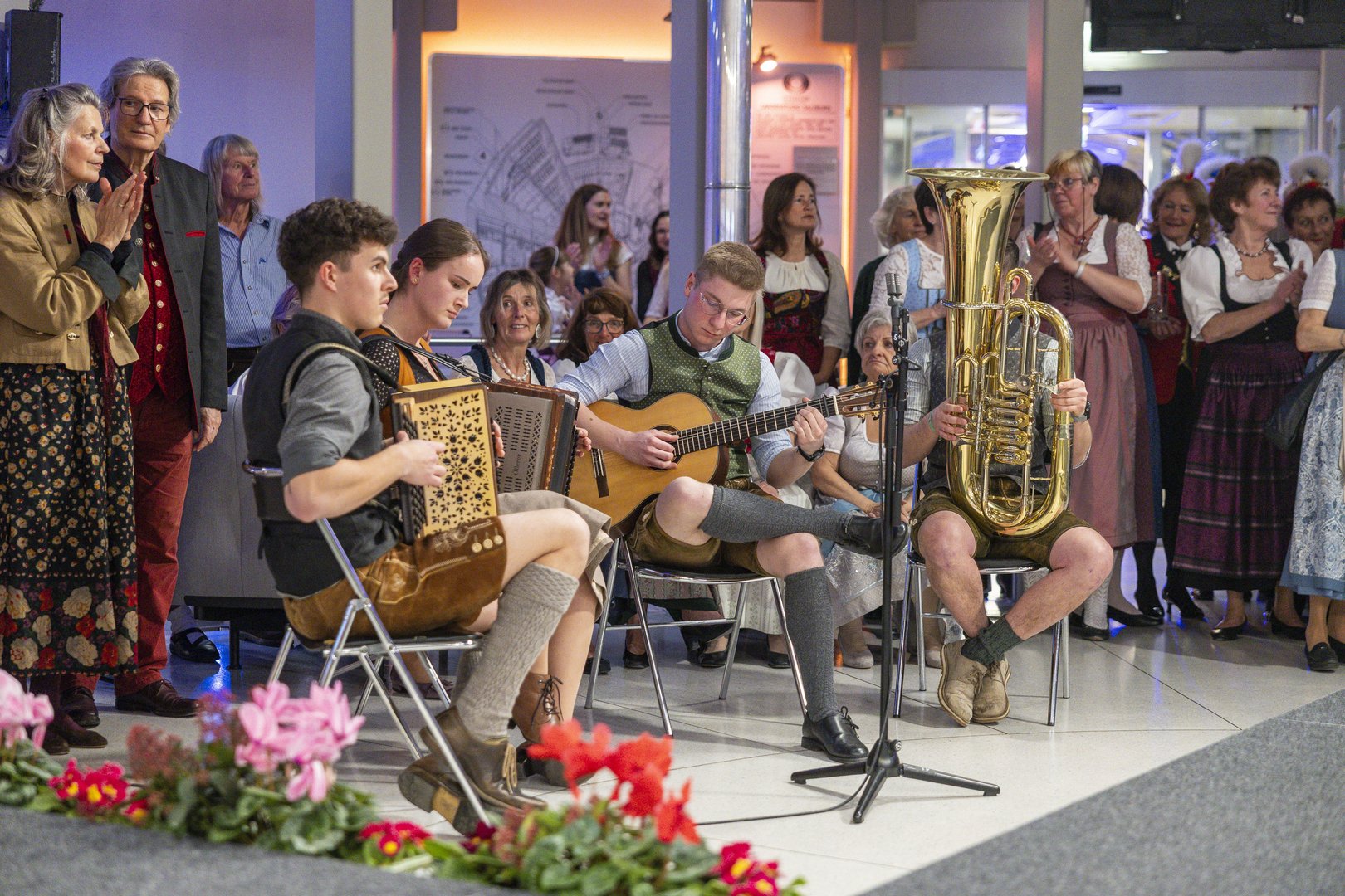 Eine Gruppe von Musikern in traditioneller Kleidung spielt vor einem Publikum in einem Innenraum, mit Blumen im Vordergrund.