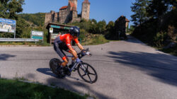 Radfahrer in Rot fährt auf einer Straße in der Nähe einer mittelalterlichen Burg unter blauem Himmel.