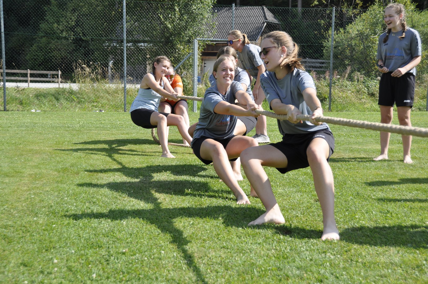 Vier M&auml;dchen in grauen Hemden spielen Tauziehen im Gras, ziehen an einem Seil und l&auml;cheln an einem sonnigen Tag im Freien.