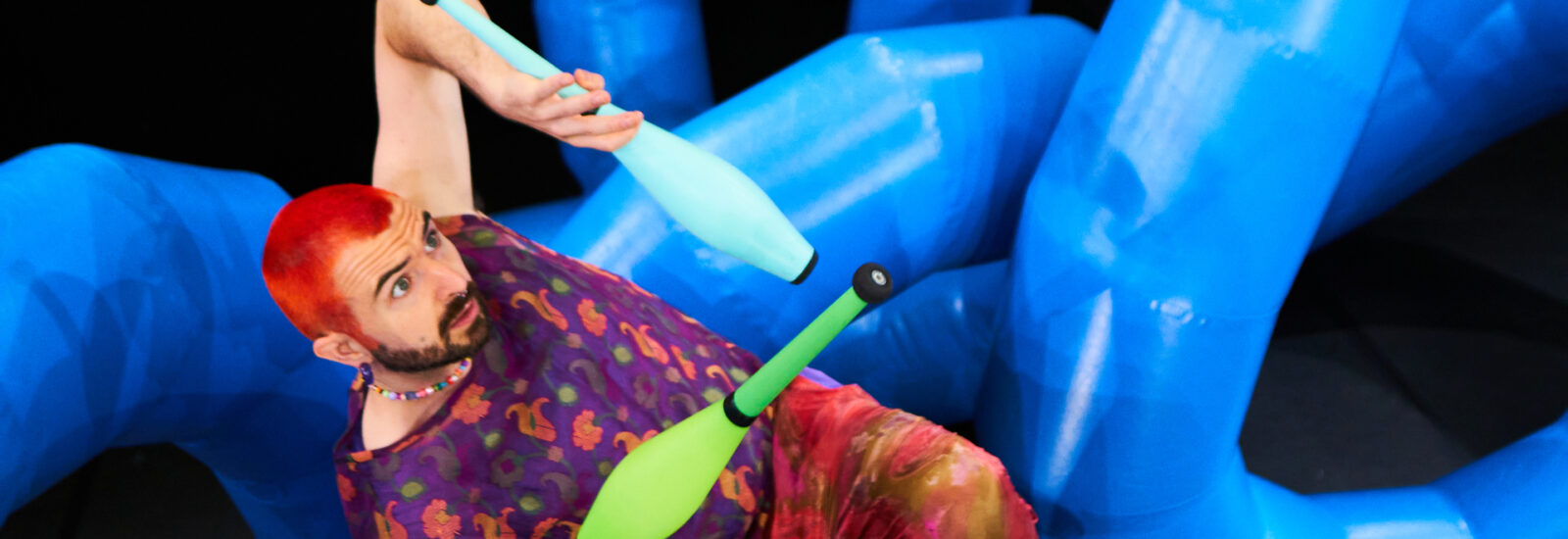 Ein Mann mit roten Haaren jongliert auf der Bühne mit bunten Keulen vor großen, sich windenden blauen Ballonfiguren.